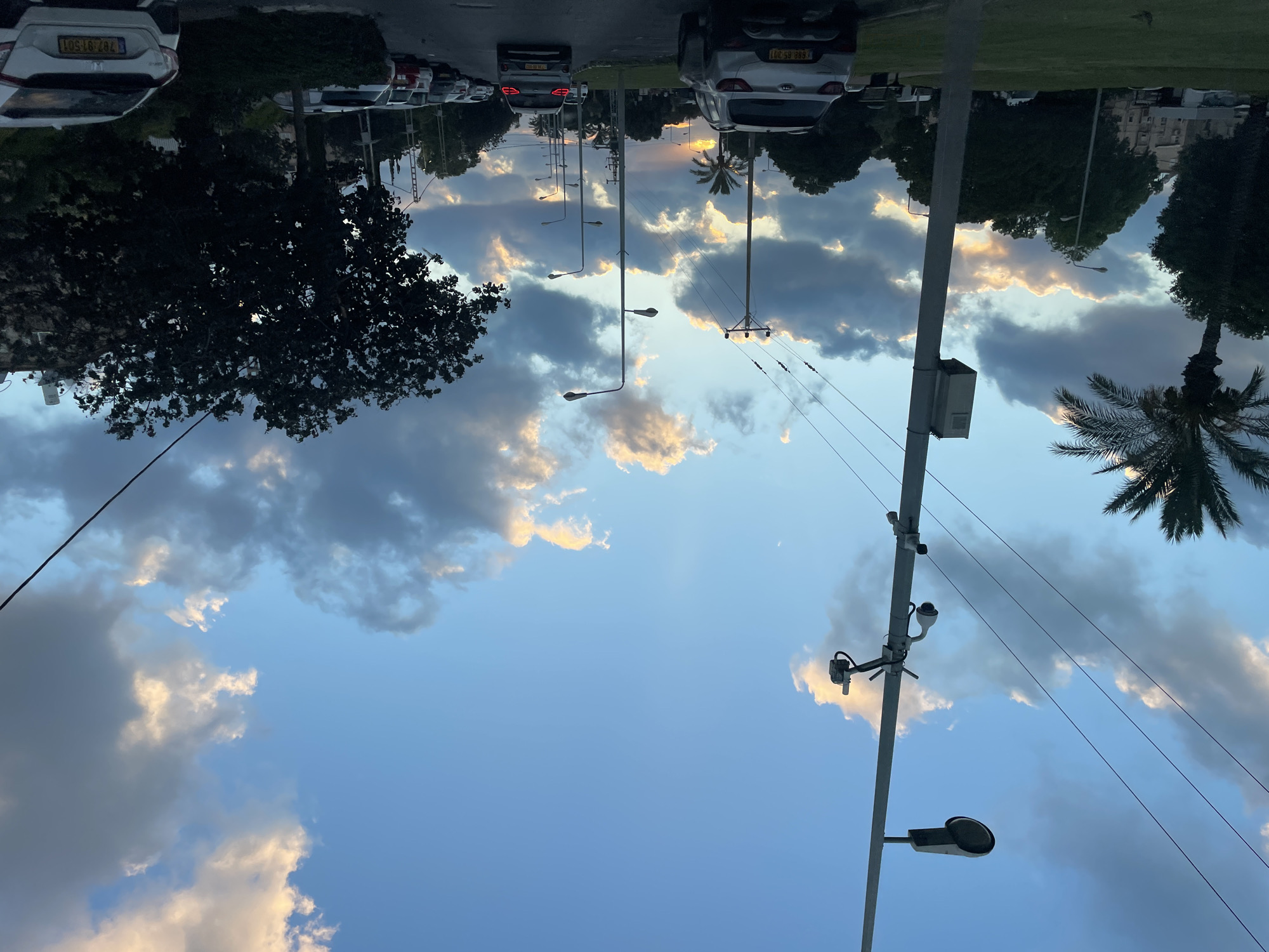 The image depicts a street scene in Haifa, Israel, during sunset. The sky is filled with dramatic clouds, illuminated by the setting sun, creating a colorful contrast with the blue sky. A tall lamppost with attached cameras and electrical equipment stands prominently on the left side of the road. Power lines stretch across the frame. Several cars are visible, parked on the side of the road and driving down the street, including a silver SUV in the foreground with license plate number "1688 85-301". Palm trees and dense foliage line the street, with buildings faintly visible in the background. The overall atmosphere is serene, capturing a moment of urban twilight.