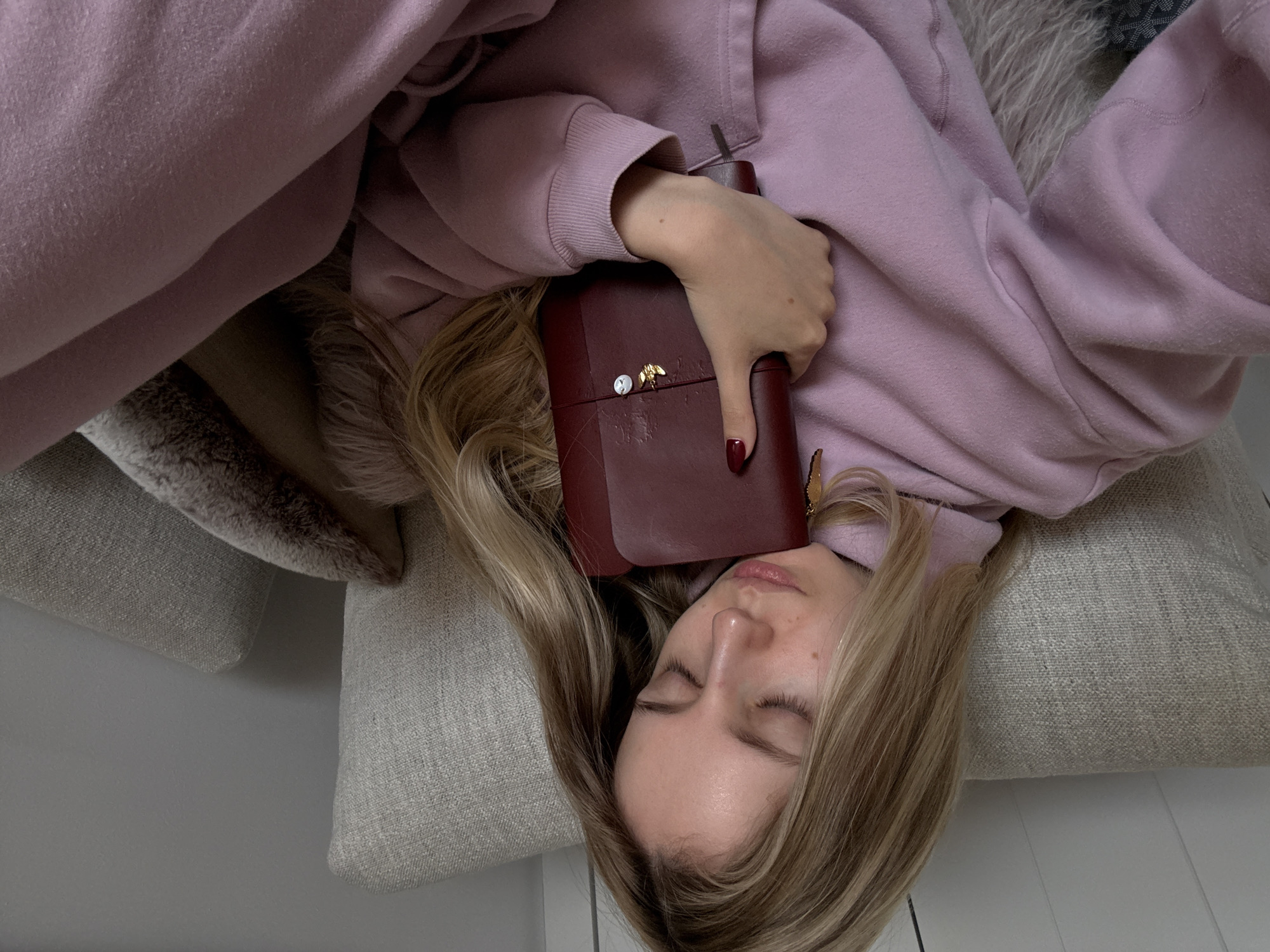 A young female subject is observed indoors in Mexico City, Mexico, positioned on a light-colored, textured upholstered couch. The subject has long, light-blonde hair and her eyes are closed, indicating a state of rest or sleep. She is wearing a light purple hooded sweatshirt and matching light purple sweatpants. Her left hand, adorned with dark red nail polish, holds a dark red rectangular object, possibly a notebook or personal organizer, against her upper torso. This object features a small gold-colored bee or insect charm and a white ribbon charm attached to its exterior. A light brown faux fur pillow is partially visible beneath her. The immediate environment includes a plain light gray wall in the background and white horizontal paneling on the upper left section. A patterned textile is visible at the bottom left of the frame.