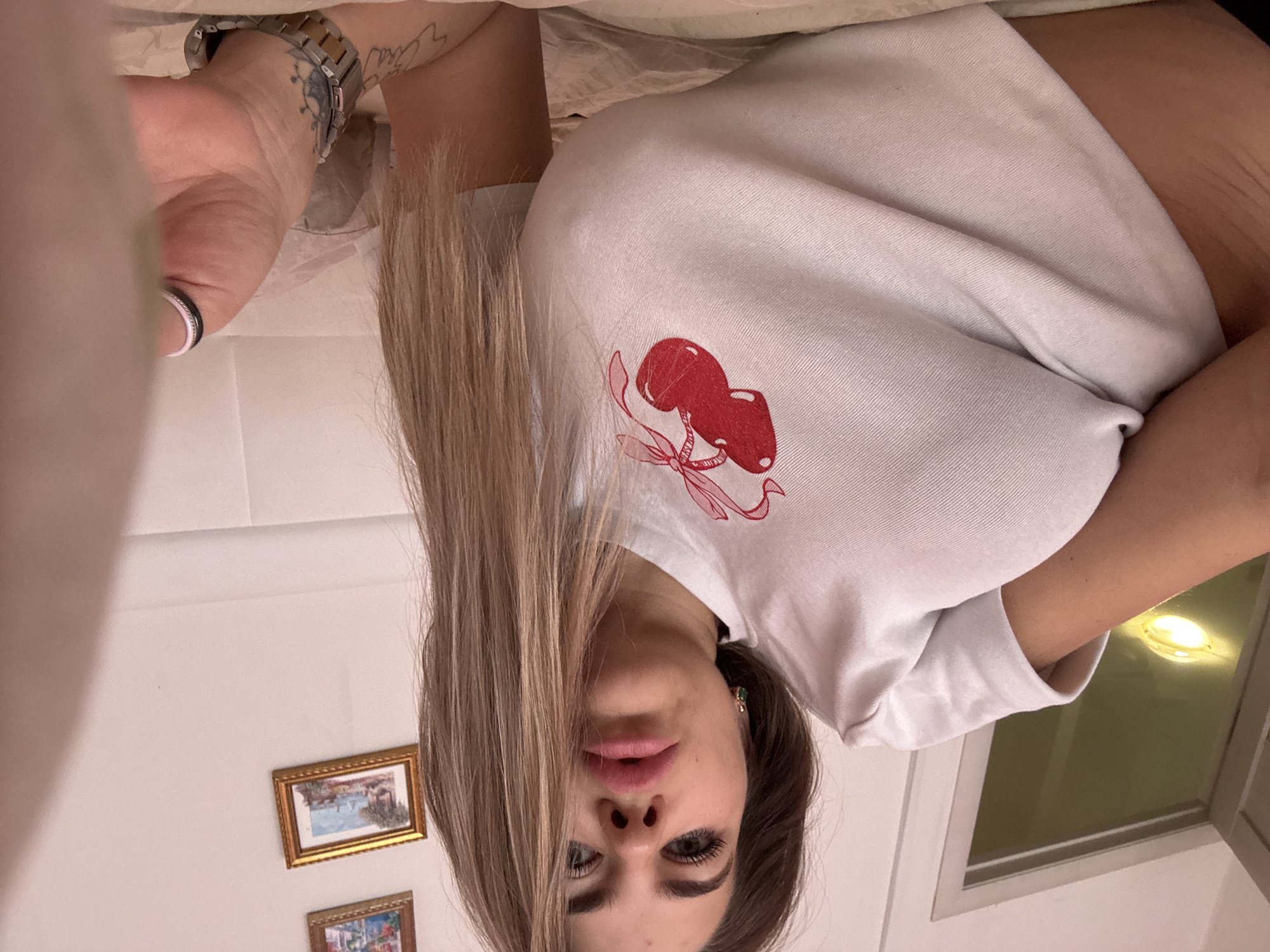 A selfie taken indoors, likely in a domestic setting. A woman with long blonde and brown hair is angled in the center of the frame. She is wearing a white t-shirt with a cherry graphic. On her left wrist, a silver-toned watch is visible, along with a ring on her finger. Behind her, two small, framed pictures are mounted on a light-colored wall. To the right, a window is observed with a light fixture reflecting within. The specific location in the city of Chaikovsky, Russia, cannot be determined from the provided visual information.