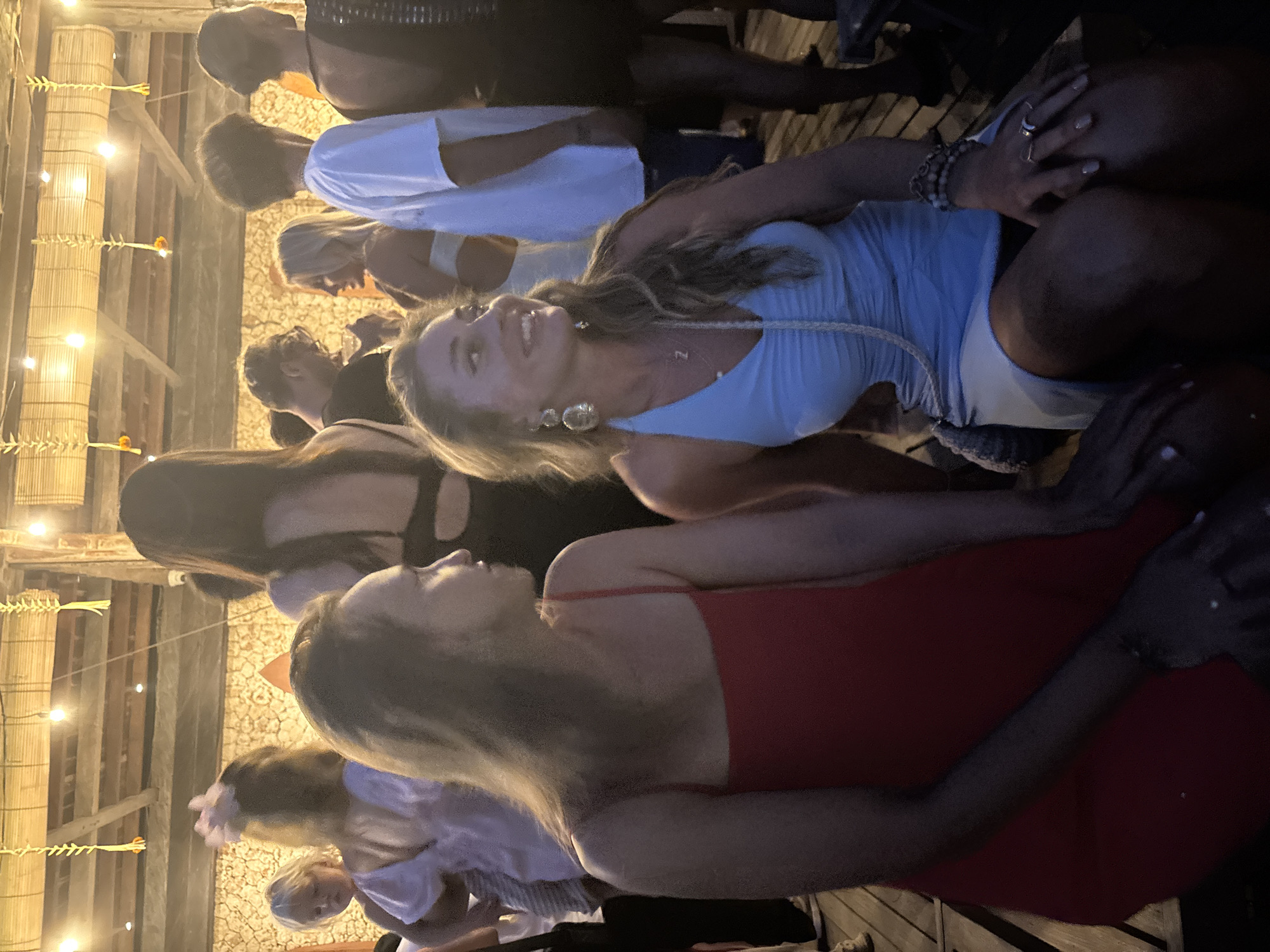 The image depicts a lively evening social gathering set in an outdoor or semi-outdoor venue, consistent with the Kuta Selatan, Indonesia location. Numerous young adults are visible, socializing under warm, ambient lighting provided by string lights strung across wooden beams and bamboo-like structures.
In the foreground, a woman with long, light brown hair, wearing a light blue dress, large silver earrings, and a necklace with a 'Z' pendant, is smiling broadly and looking upwards, conveying a joyful expression. Her hands, adorned with rings and bracelets, are visible. Next to her, another woman with light hair is seen from behind, dressed in a red top or dress, with her arm resting on a surface. Other individuals, some wearing white shirts or dark clothing, are present in the background, partially obscured but contributing to the active scene.
The setting features rustic architectural elements such as wooden beams and what appear to be bamboo panels or woven material. Decorative elements, possibly made from woven leaves or dried plant material with small orange flowers, hang near the string lights, adding to the tropical or Balinese aesthetic. The ground beneath the subjects appears to be wooden flooring or decking. The soft, artificial lighting indicates that the event is taking place at night or during the evening. The overall mood is social, relaxed, and cheerful.