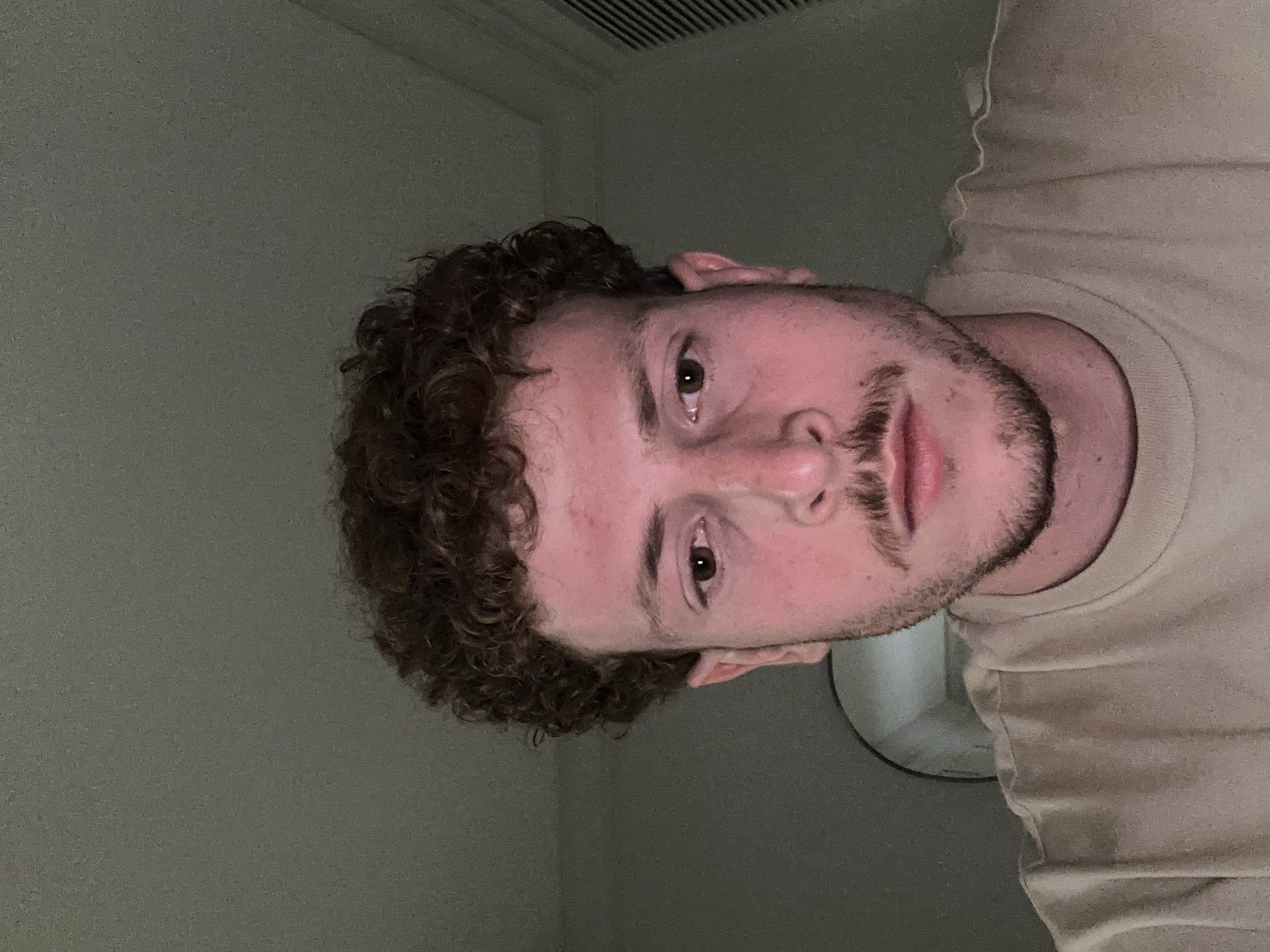 A medium, indoor shot shows a fair-skinned male facing slightly right. His head is tilted to his right, so his left eye is higher than his right. He has short, brown, curly hair and a short, brown beard. He is wearing a tan t-shirt. The background includes an off-white wall, a louvered ceiling vent, and what appears to be a round fixture. This image does not provide information regarding the city of Aventura, United States of America.