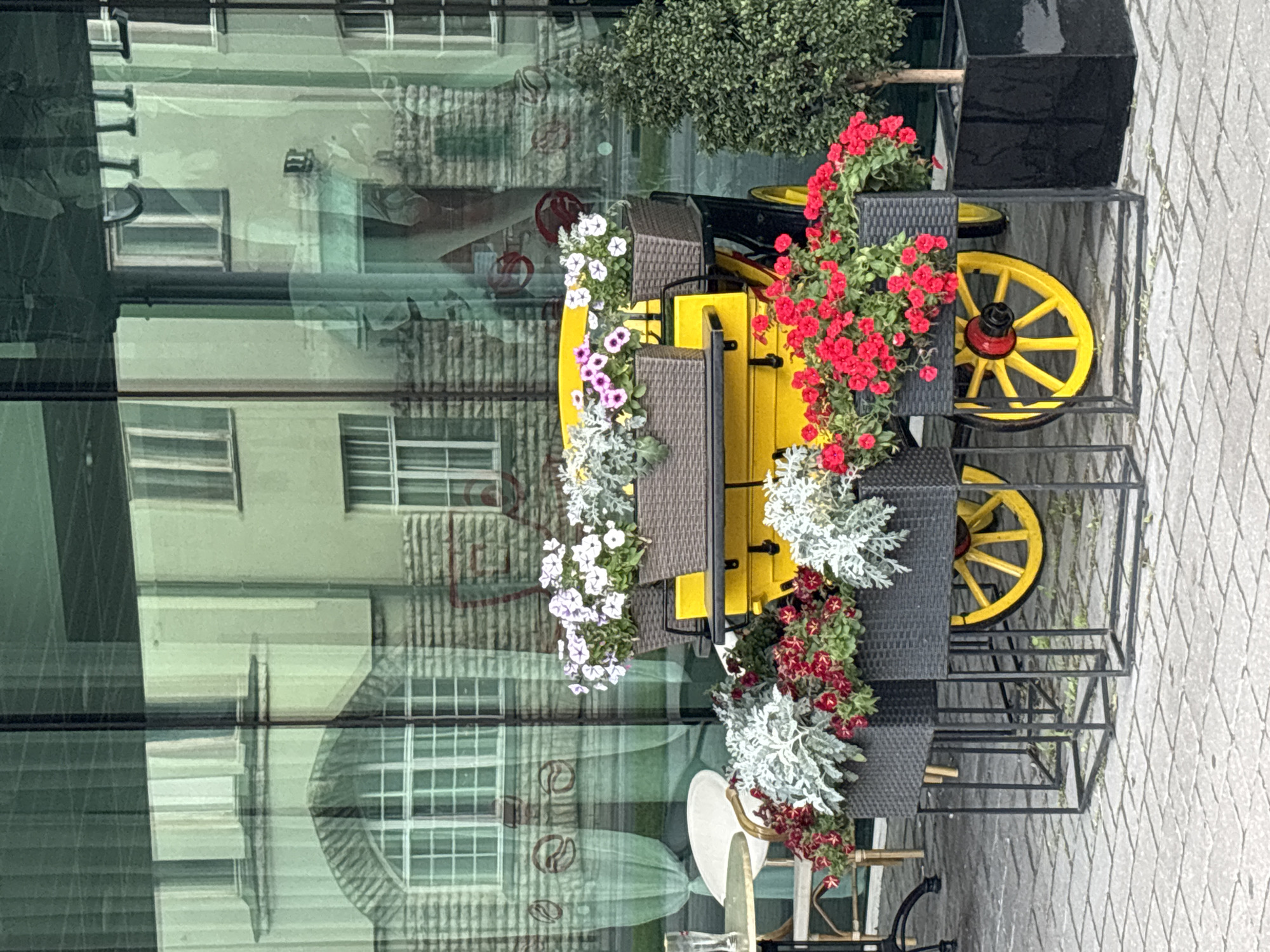 The image depicts a street scene in Tallinn, Estonia, with a yellow decorative carriage adorned with flowers. The carriage is positioned in front of a building with large glass windows, reflecting the architecture of the surrounding structures. In the foreground, several planters filled with red and white flowers and silver-grey foliage are arranged on a brick-paved sidewalk. To the right of the carriage, a small, manicured tree in a black planter stands beside the building. In the background, outdoor seating for a cafe or restaurant is visible, including a round table and chairs, partially obscured by the carriage and planters. The overall scene suggests a pleasant outdoor setting, likely in a public area or near a commercial establishment in Tallinn.