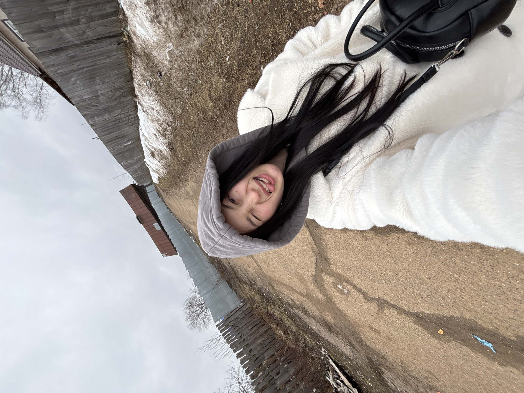 The image shows a woman with long black hair wearing a gray hooded sweatshirt and a white, fluffy jacket. She is smiling with one eye closed, seemingly taking a selfie. A black bag with a handle and strap is visible near her. She is outside on a dirt road or path with a wooden fence on one side and patches of snow on the other. The sky is overcast. Based on available information, the location appears to be in the city of Фоки, Russia.