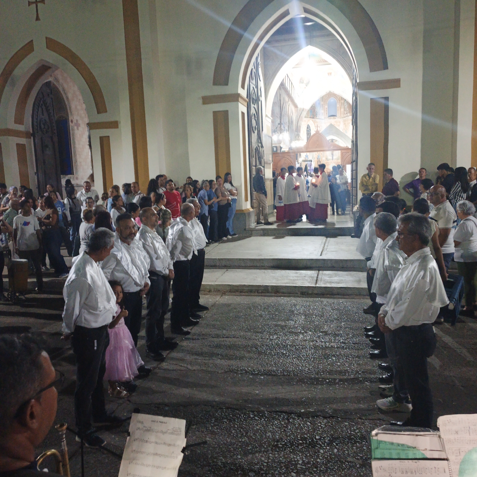 The image depicts a large group of people gathered outside a church. Many people are standing in orderly lines, some wearing white shirts and black pants, while others are in the background wearing a variety of clothing. There are religious figures wearing white robes with red accents, holding candles. The church has a prominent arched entrance. In the foreground, a musician with a trombone stands with music sheets. The setting is illuminated, and the overall scene suggests a religious procession or ceremony is taking place. Location: Tinaquillo, Venezuela.