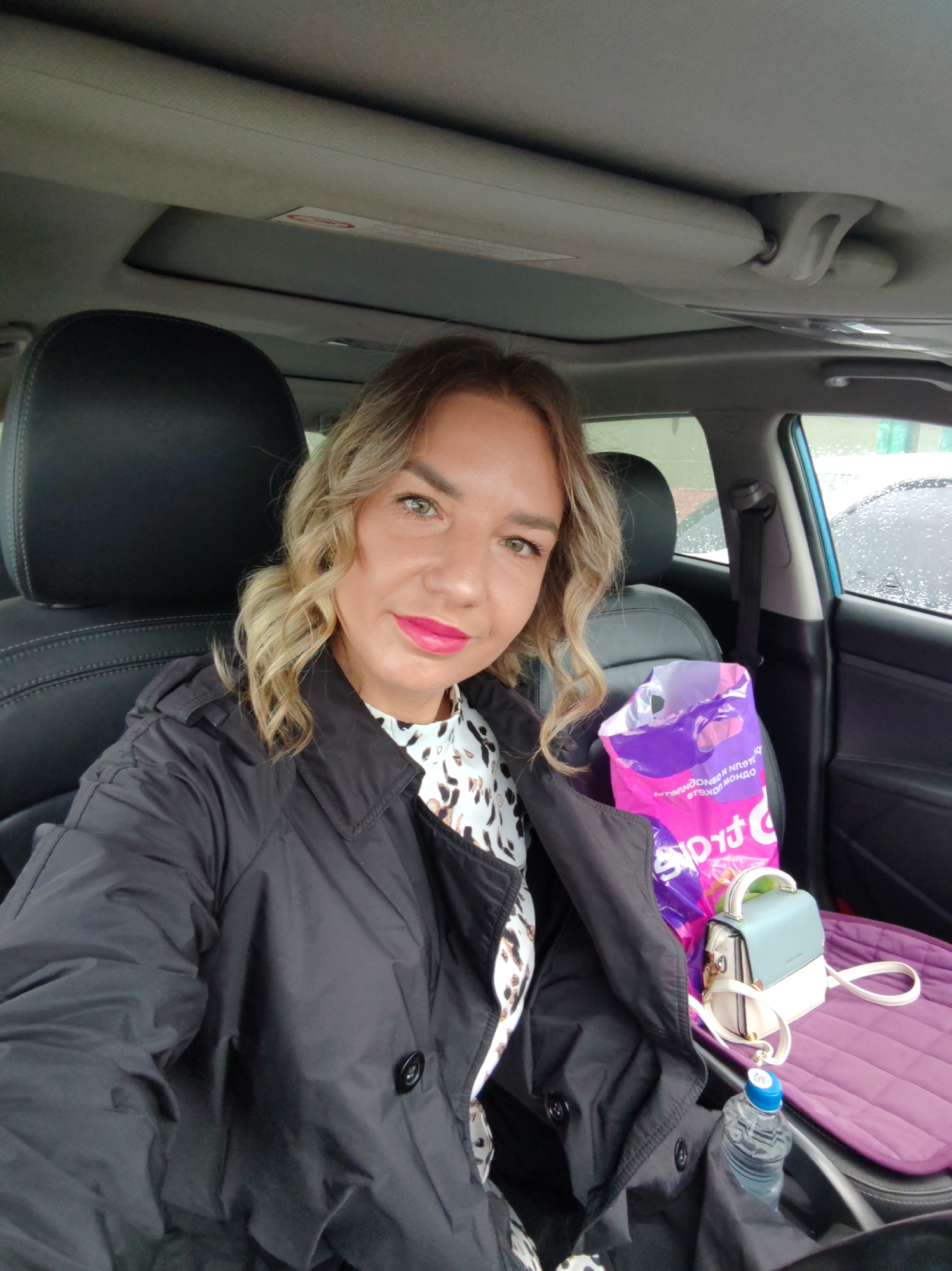 A woman is sitting in the driver's seat of a car. She is wearing a black coat over a patterned blouse and has blonde, wavy hair. She has pink lipstick. The car has black leather seats and a sunroof. On the passenger seat is a purple shopping bag, a light blue and white purse, a purple quilted seat cover, and a bottle of water. Outside the car, there are other cars and a building visible. The city of Petropavlovsk-Kamchatsky, Russia is not discernible from the image. The weather appears to be overcast.