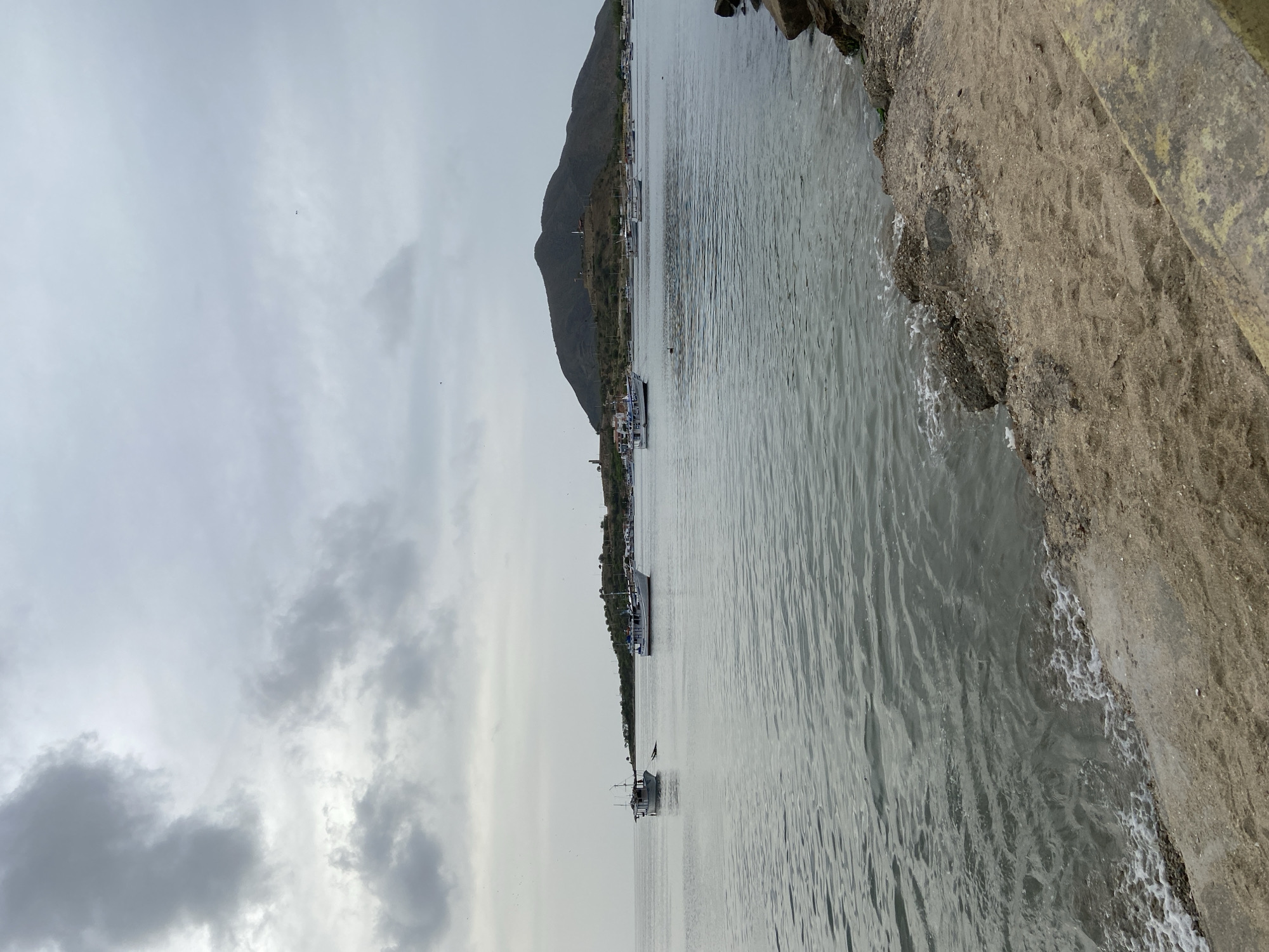 This image depicts a cloudy day at the waterfront in Juan Griego, Venezuela. The foreground shows a sandy and rocky shoreline. The body of water in the middle ground is calm with gentle waves lapping the shore. Several boats are visible on the water, ranging from small fishing vessels to larger ships. In the background, a hill rises above the coastline, dotted with buildings and structures. The overall atmosphere is overcast and serene.