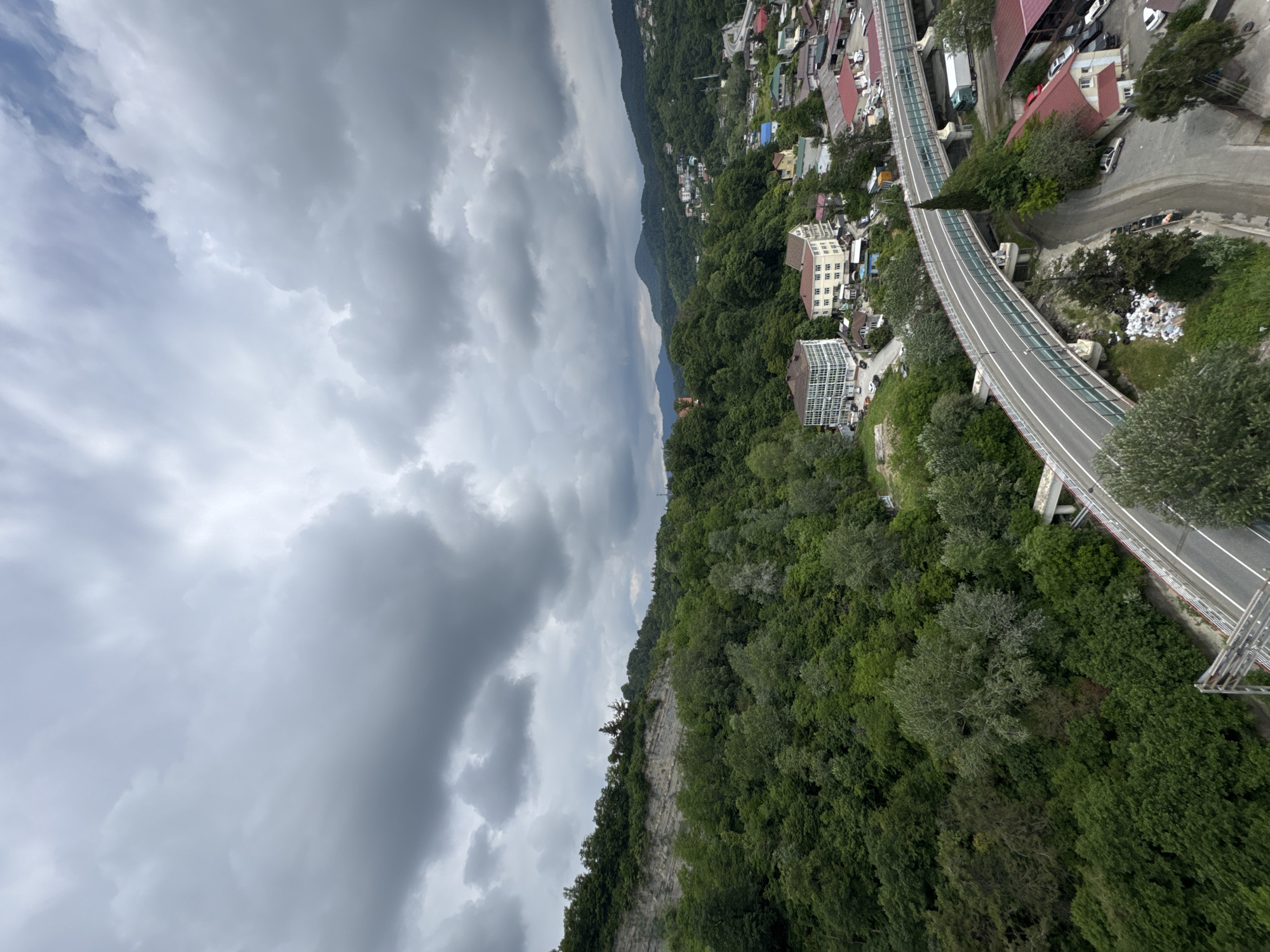 The image is a high-angle, wide shot of a coastal city in Sochi, Russia, featuring a dramatic, overcast sky filled with dark, textured clouds. In the foreground, a multi-lane highway bridge curves through the scene, with white guardrails and a glass barrier along its length. Beneath the bridge, the landscape transitions from lush green trees on a rocky slope to a more developed area with buildings, red-roofed structures, and parked vehicles.
Further into the frame, the city sprawls across rolling, densely forested hills. Several multi-story buildings with white and beige facades are visible, some appearing to be hotels or apartment complexes. Numerous smaller residential buildings with red roofs are scattered throughout the landscape, indicating a residential area. The hills are covered in a dense canopy of green trees, with some appearing lighter in color, suggesting a mix of deciduous and evergreen species. The distant mountains are shrouded in a hazy, green blanket. A white truck is present on a road adjacent to the bridge. Scattered debris is visible on the ground on the left side of the bridge. The overall impression is one of a city nestled within a heavily vegetated, mountainous terrain under a moody sky.