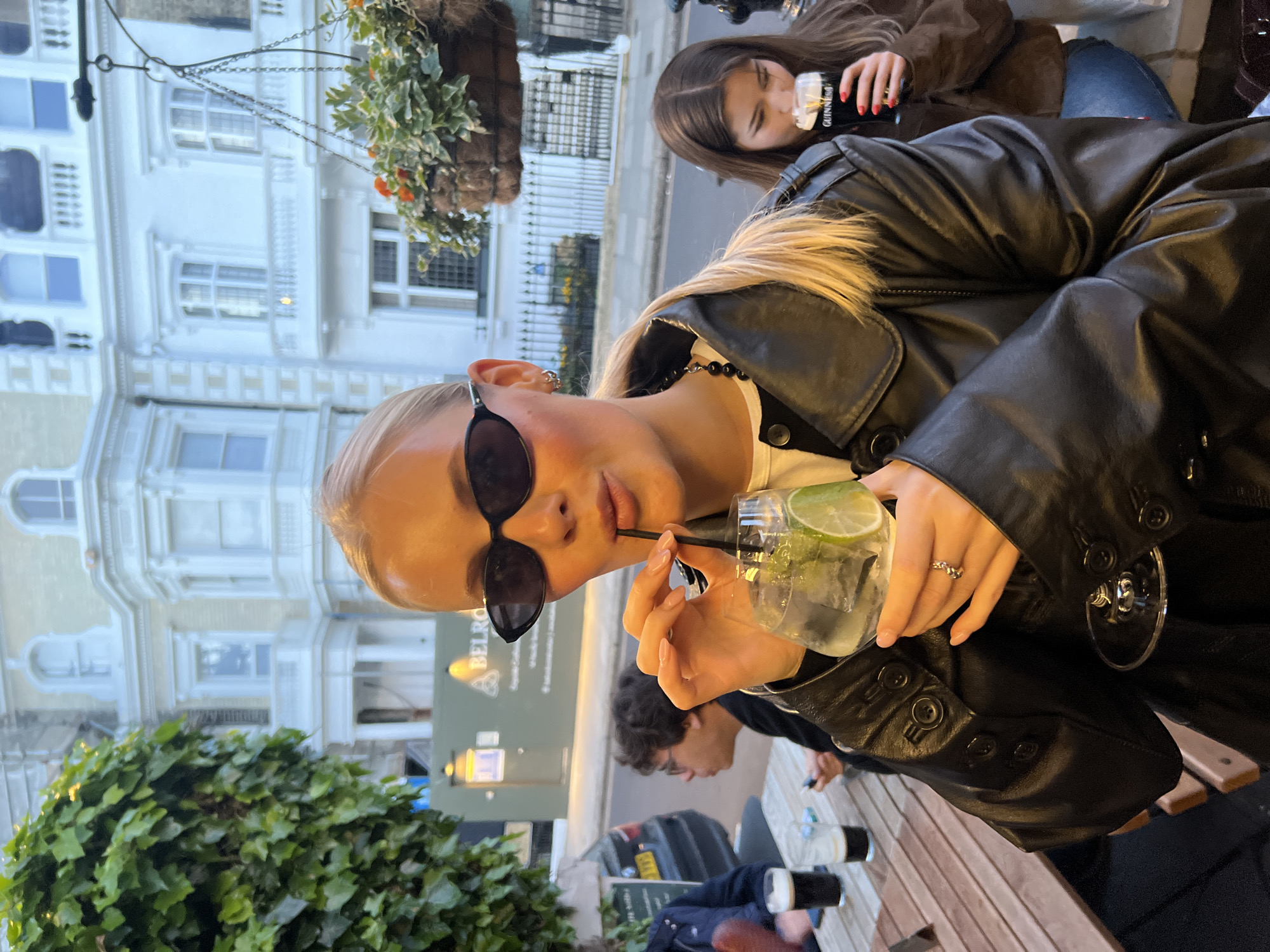 A medium, eye-level shot depicts a group of people at an outdoor cafe table. A young, blonde woman in a black leather jacket is holding a glass of water with lime and a straw and looking at the camera. She is wearing sunglasses and a black beaded necklace. Another woman with dark hair is in the background, holding a bottle. A man with dark hair is also at the table. Several glasses and bottles are on the table. The background shows a building with white windows and ornate details. A hanging basket with greenery is visible above. A sign indicates this is the "Belrose," suggesting the location is in London, United Kingdom.