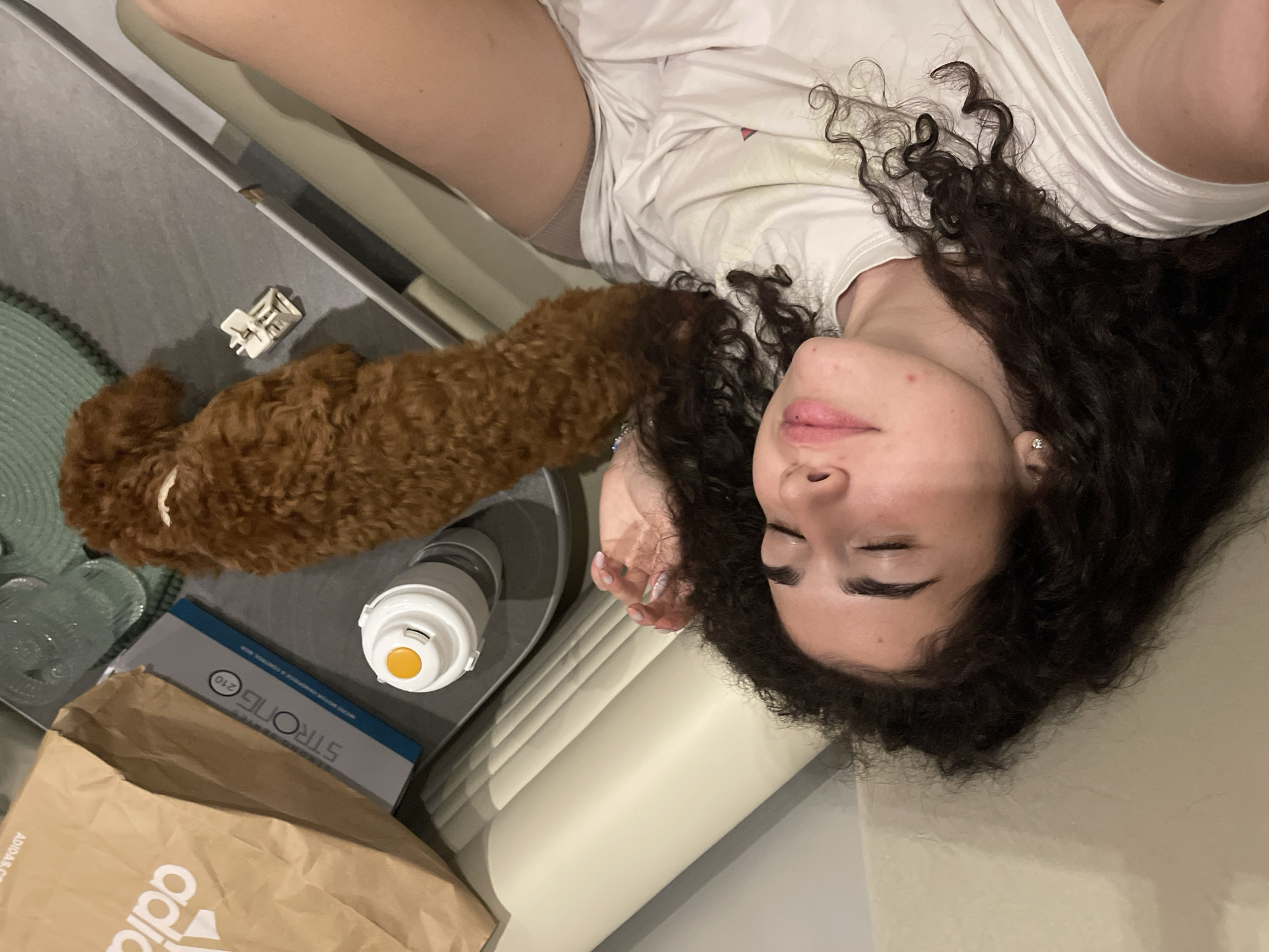 An individual with dark, curly hair is lying on a light-colored upholstered surface, appearing relaxed with closed eyes or looking upwards. They are wearing a white t-shirt, and a small stud earring is visible in their left ear. Their left hand, showing light nail polish, rests near their head. Adjacent to the individual, on a grey flat surface, a small, reddish-brown, curly-haired dog with a light-colored band around its neck is also lying down.
To the left of the scene, a brown paper bag with a partial "adidas" logo is visible, alongside a rectangular package labeled "STRONG 210" and some smaller text below. A white and grey thermos or water bottle with an orange top is positioned in front of the package. A small, white, plastic, multi-pronged object lies on the grey surface near the dog. A textured green mat or item is partially visible in the background. The overall environment is an indoor, domestic setting, likely a living room or bedroom. The image provides no identifying information regarding a specific city or country.