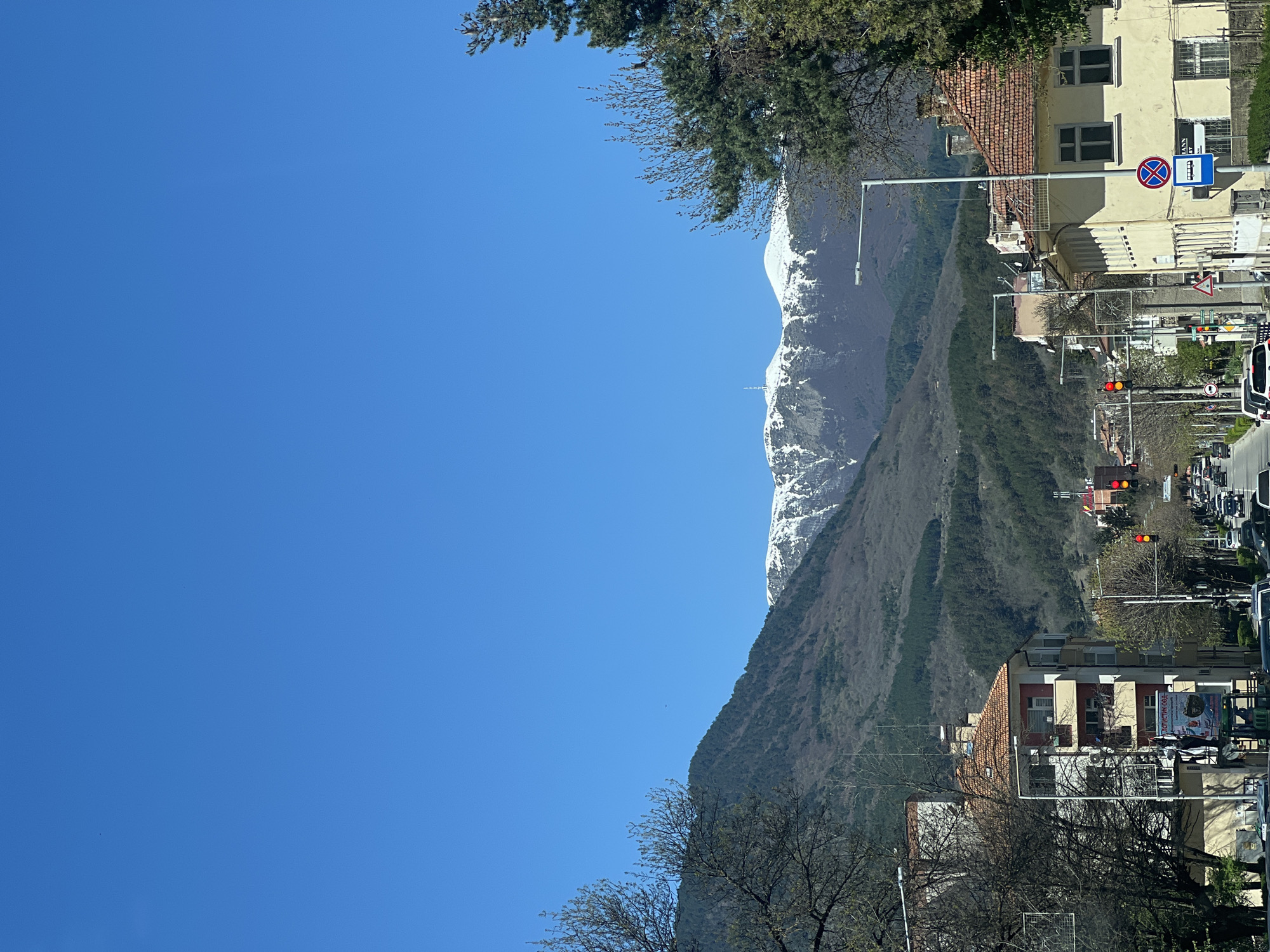 A vertically-oriented outdoor shot shows a street in Goce Delcev, Bulgaria, with a clear blue sky occupying much of the left side. A snow-capped mountain range is visible in the background, partially obscured by a green, tree-covered mountain slope. On the right side of the frame, a typical European city street is visible, including buildings with red-tiled roofs, street lights, a bus stop sign, traffic lights showing red, cars, and trees.