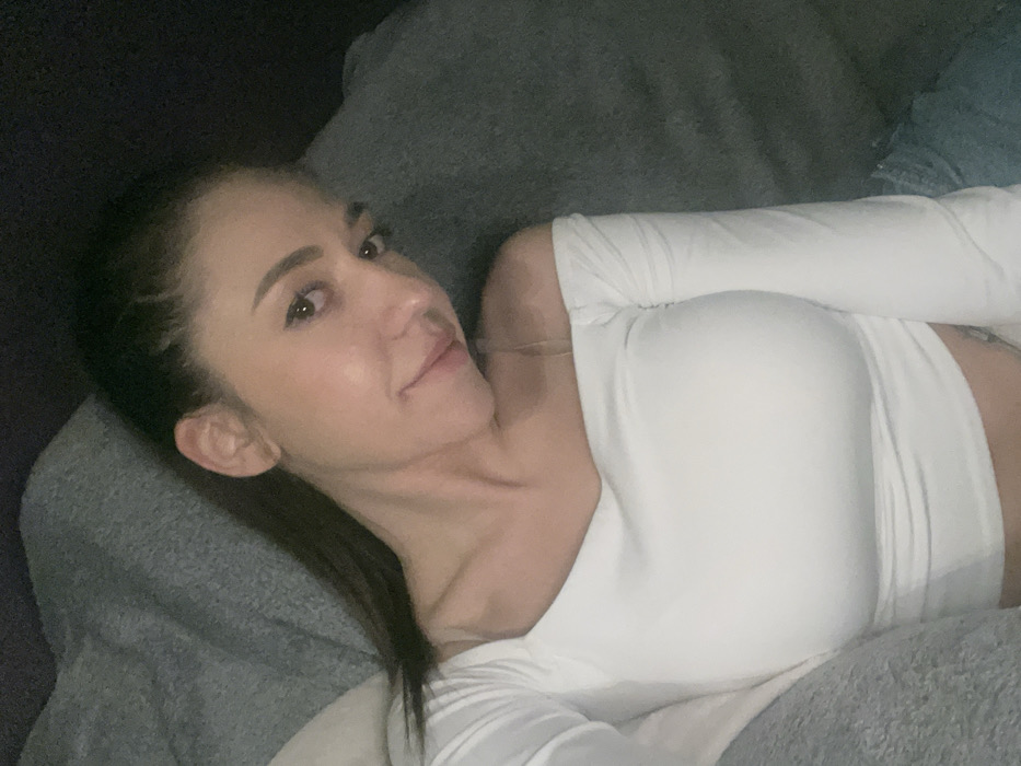 A single female individual with dark hair is depicted in a medium-close-up shot, appearing to be a selfie taken indoors. She is positioned reclining against a soft, grey-toned fabric background, consistent with an upholstered piece of furniture such as a couch or bed. The subject has fair skin and is looking directly at the camera with a slight smile. She wears a white, form-fitting, long-sleeved, off-the-shoulder top with a wide neckline, exposing her shoulders and a portion of her midriff. A thin, light-colored strap is visible on her left shoulder. Part of a light-colored garment, possibly denim, is visible near her lower right. The lighting is soft and somewhat dim. The specific location within the city of Sofia, Bulgaria, cannot be determined from the visual information presented.