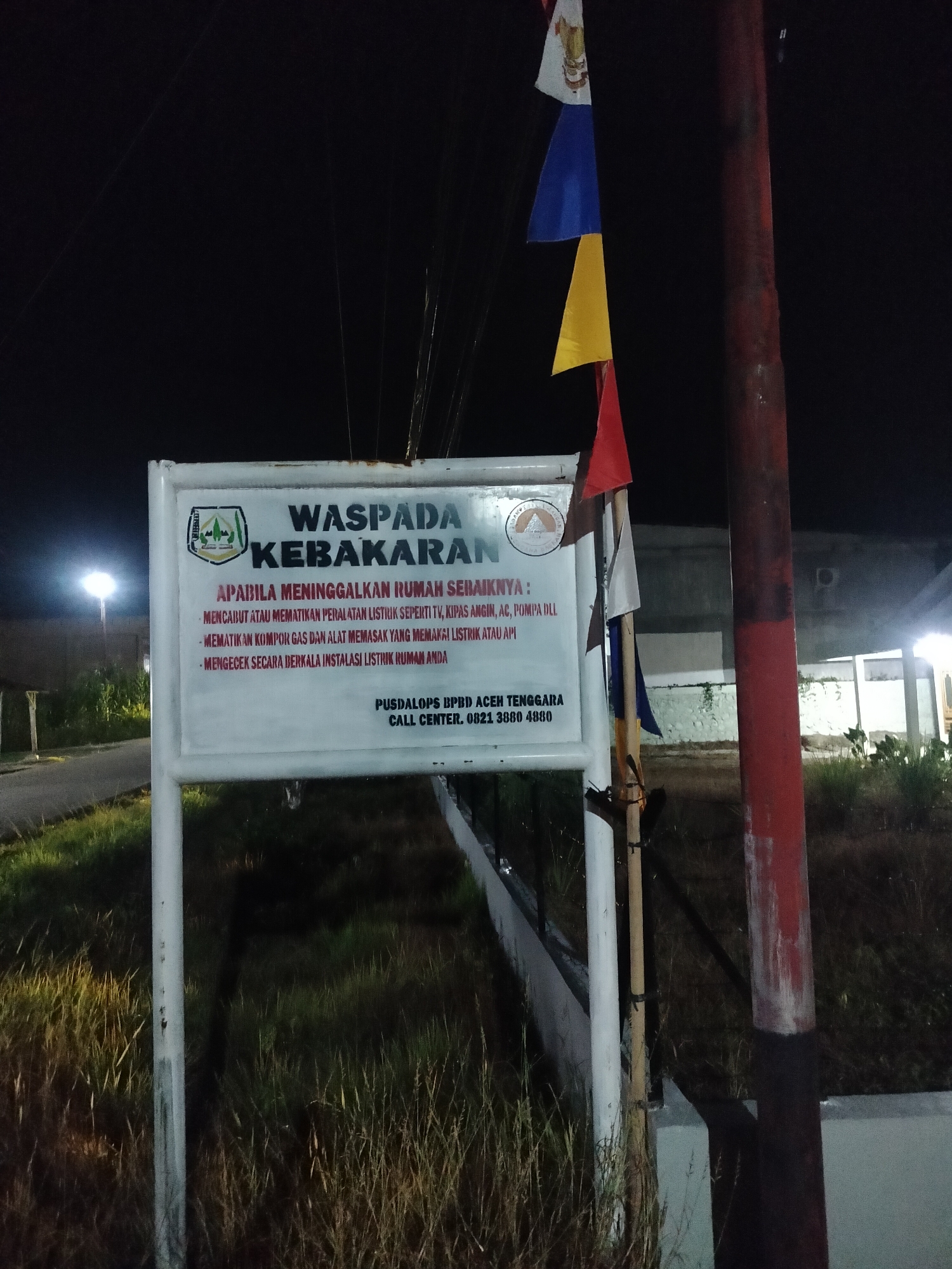 In Kumbangindah, Indonesia, a sign with red and white framing is positioned in front of a grassy area at night. The sign features text in Indonesian, with the prominent header "WASPADA KEBAKARAN" (Fire Alert). Below this, it provides instructions on what to do when leaving a house, such as unplugging electrical appliances like TVs and air conditioners, turning off gas stoves, and regularly checking electrical installations. The sign also includes contact information for "PUSDALOPS BPBD ACEH TENGGARA" and a call center number. Three flags, white, blue, and yellow, are attached to a pole near the sign. In the background, a building with illuminated windows is visible, and a red pole stands to the right of the sign. A lamppost can be seen in the distance to the left, casting light on the scene.