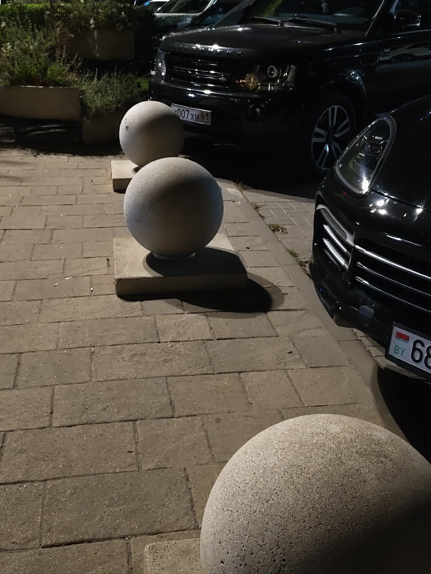 The image captures a nighttime street scene in Rostov-on-Don, Russia. The foreground is dominated by large, spherical concrete bollards, strategically placed along a cobblestone sidewalk. These bollards, designed to prevent vehicle access, are lit by an unseen light source, casting shadows on the ground. In the background, several black luxury SUVs are parked. One SUV, a Range Rover, has its license plate partially visible, displaying Cyrillic characters and numbers. Another vehicle, a Porsche Cayenne, is also present, its distinctive headlights and grille visible. The overall impression is one of a well-maintained urban environment, likely a commercial or residential area, where security measures are in place to manage vehicle traffic.