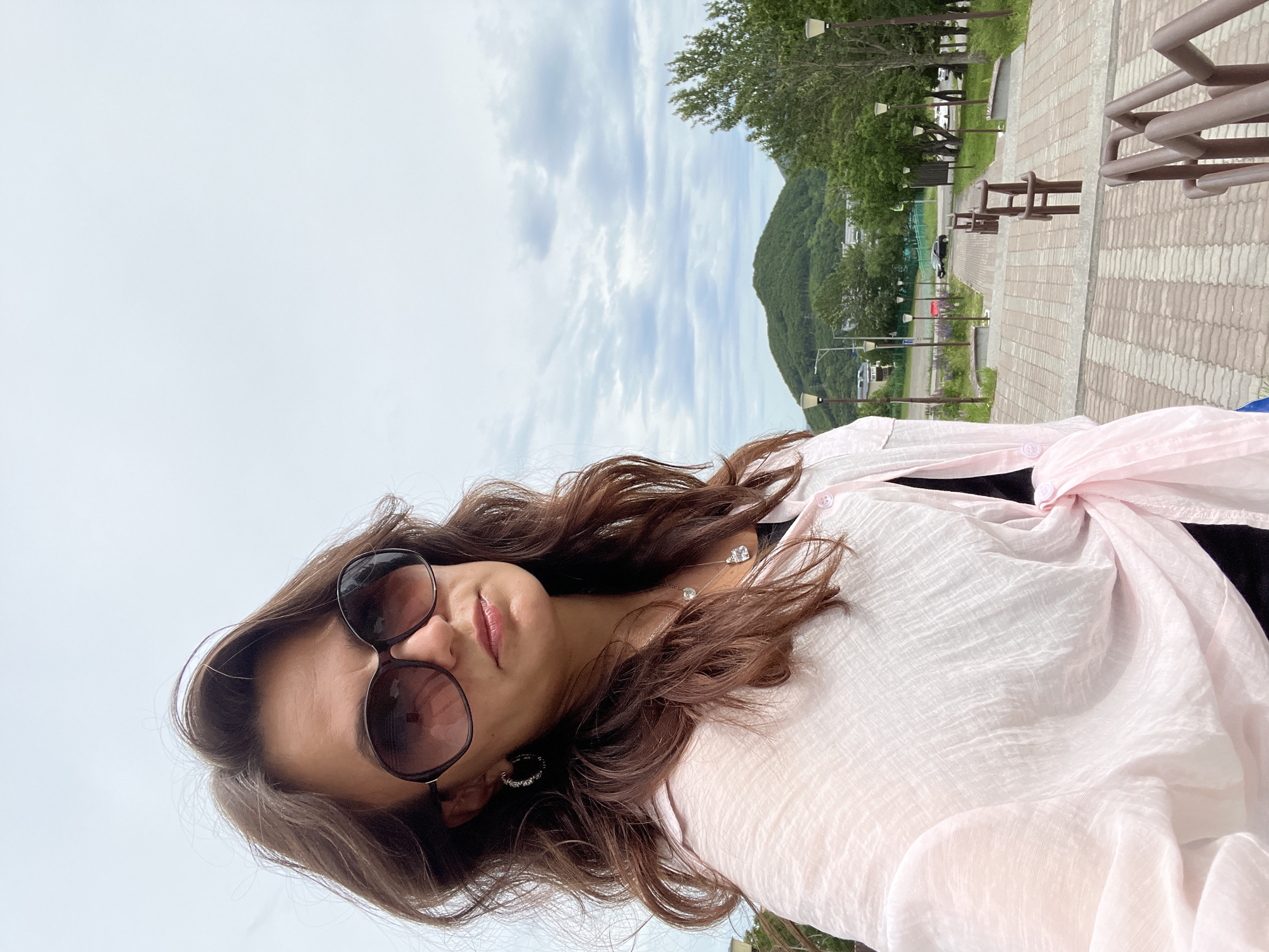 A medium shot captures a woman with long, wavy brown hair and large sunglasses taking a selfie. She is wearing a sheer pink button-up shirt, partially unbuttoned, revealing a black garment underneath. A silver pendant necklace with a large clear stone hangs around her neck, and she wears hoop earrings. Her hair is styled loosely, framing her face.
In the background, a wide paved walkway with brick-like patterns descends a gentle slope. Lampposts line both sides of the walkway, and there are metal railings. Beyond the walkway, a green landscape unfolds, featuring trees and a prominent, rounded, tree-covered mountain under a cloudy sky. The setting appears to be an outdoor public space, likely a park or scenic overlook, in the city of Petropavlovsk-Kamchatsky, Russia, given the characteristic mountainous terrain and potentially the architecture of the distant buildings. The overall atmosphere is casual and outdoor-oriented.