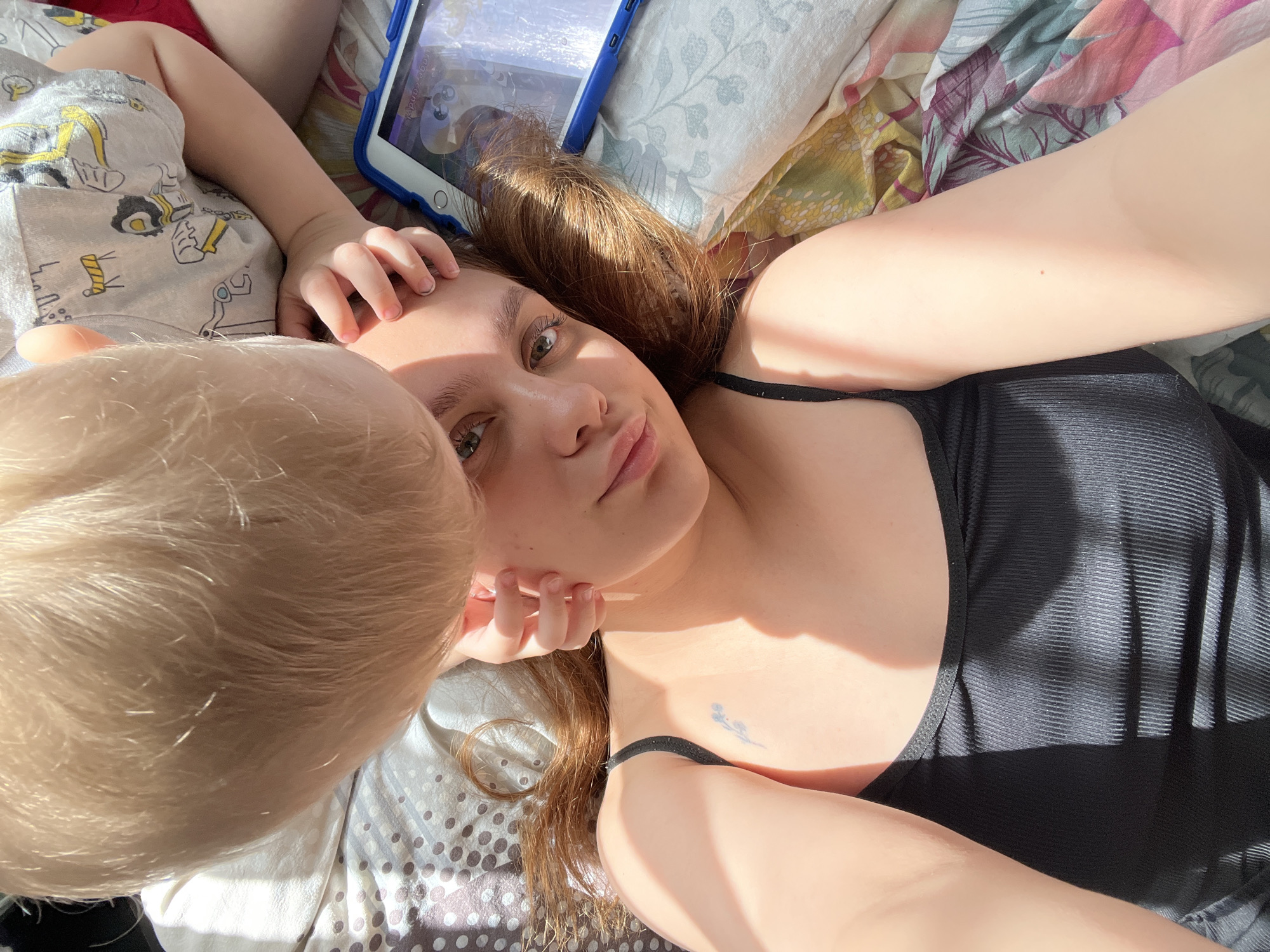 A woman lies on a bed with two children. The woman is light-skinned with auburn hair and green eyes. She wears a black tank top and a black elastic hair tie on her wrist. She has a small, blue tattoo on her chest. Her head is resting on her hand. A child with blonde hair is close to her face. Another child with light skin wears pajamas printed with construction vehicles. There is an iPad in a blue case lying on the bed next to the woman. The bed has floral and polka-dot sheets. The scene appears to be indoors, lit by sunlight. There is no indication of the location being Ольховка, Russia, in the image.