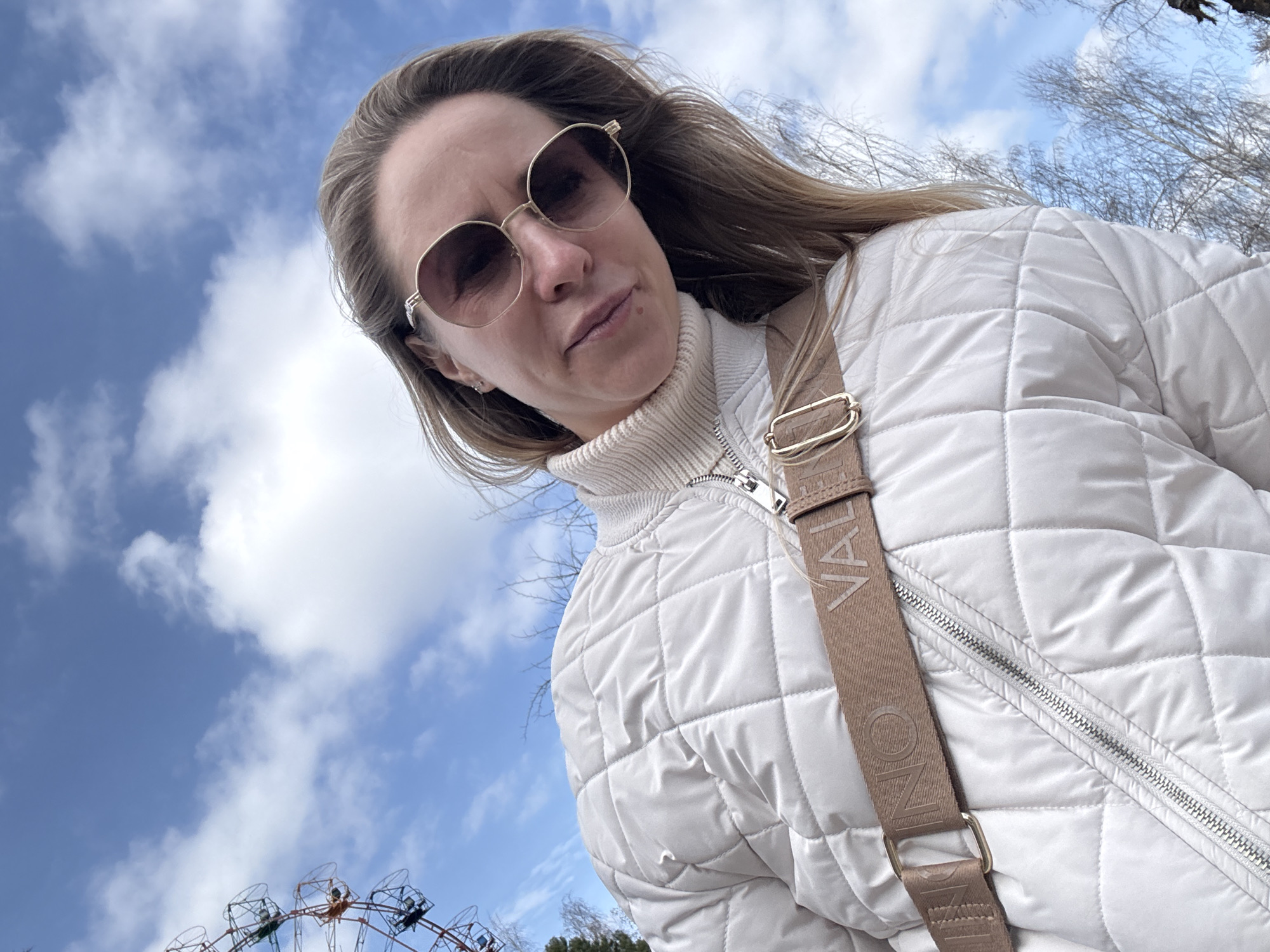 A woman is looking down at the camera, wearing sunglasses, a white quilted jacket, and a turtleneck sweater. She has a beige shoulder bag slung across her body. In the background is a blue sky with scattered white clouds. In the lower left corner of the image, a Ferris wheel is visible. The Ferris wheel indicates the location is likely in an amusement park in Chaikovsky, Russia. Trees with bare branches are also visible.