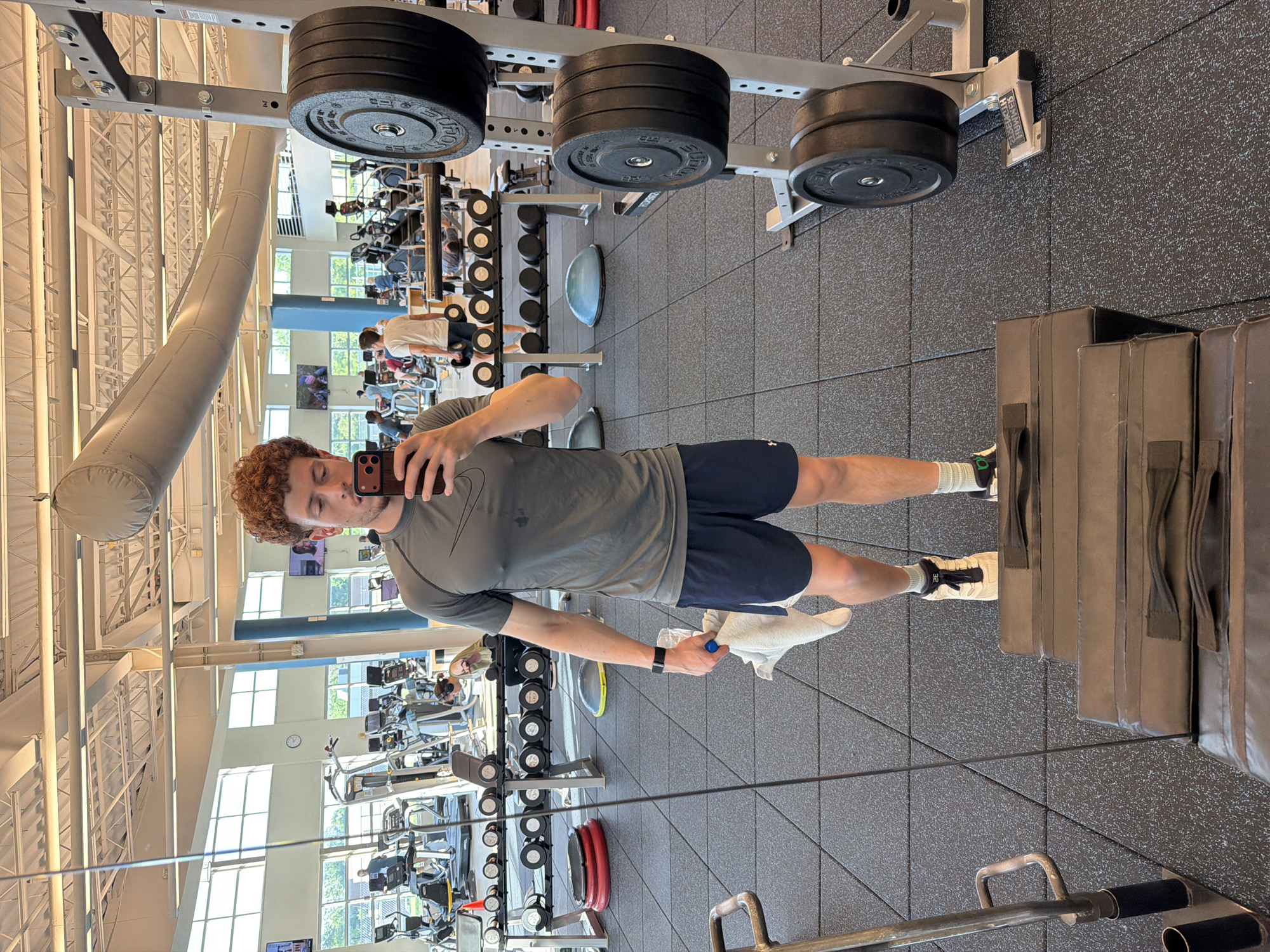 This image, rotated 90 degrees clockwise, captures a mirror selfie taken inside a spacious, modern gym located in Beachwood, United States of America. The primary subject is a young man with curly reddish-brown hair, shown from the chest down. He is dressed in a gray long-sleeved athletic shirt bearing a white Nike logo, dark blue athletic shorts, and white and green athletic shoes with the number "28" visible. He holds a dark-colored smartphone to take the photo, partially obscuring his face. A white towel is draped over his left hand, and a smartwatch is visible on his left wrist.
The gym floor is covered with a speckled black rubberized material. The facility features high ceilings with exposed white structural beams and ductwork. Large windows allow ample natural light into the space, suggesting it is daytime. In the background, reflected in the mirror, several other individuals are visible using various exercise machines and weight equipment.
Notable objects include heavy black weight plates, some branded "ROGUE," stacked on a metal rack. A metal bracket on the wall displays the text "HAMMER STRENGTH." Further back, dumbbell racks and a variety of cardio and strength training machines are visible. Padded wall mats are situated near the bottom right of the frame. The overall scene depicts a functional and busy fitness environment.