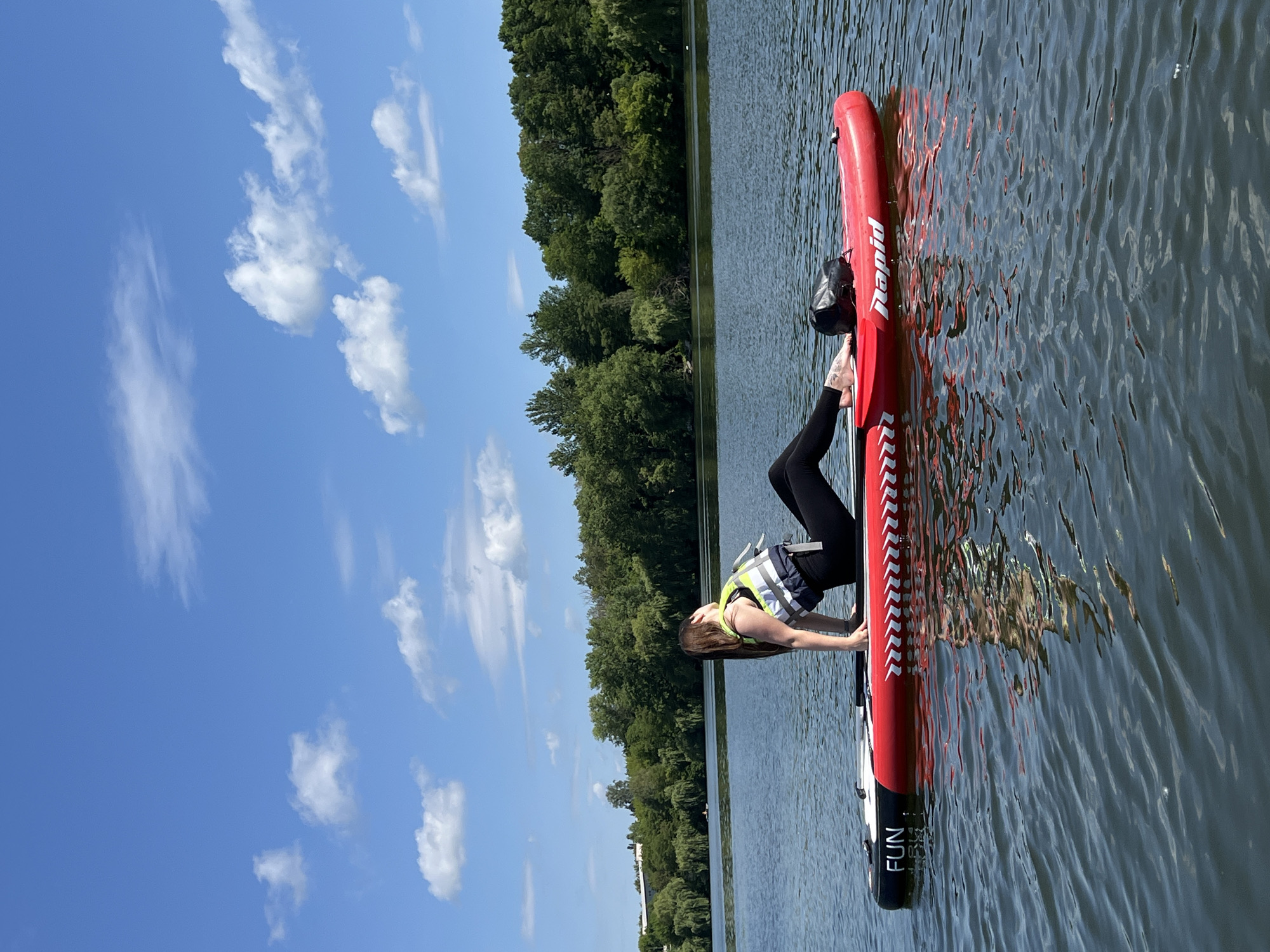 A woman is practicing yoga on a stand-up paddleboard on a lake under a blue sky with scattered clouds. The woman is wearing a life vest, black leggings, and a yellow top. The paddleboard is red and white with the words "FUN" and "Rapid" printed on it. The lake is surrounded by a line of trees and in the distance, there are buildings. The overall scene is calm and serene.