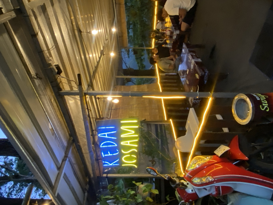 The image displays an illuminated outdoor or semi-outdoor eating establishment identified by a blue neon sign reading "KEDAI" above a yellow neon sign reading "JOSAMI." The location is within the city of Cembor, Indonesia, during evening hours, indicated by the artificial lighting and dark background.

The establishment is covered by a corrugated metal roof supported by metal poles. Warm white string lights are strung horizontally along a wooden railing, and bare light bulbs hang from the roof structure, providing primary illumination. A bamboo-style screen is visible behind the neon signage.

In the foreground, to the left, a red and white scooter (consistent with a Honda Vario model) is parked, facing the left side of the frame. Adjacent to the scooter is a white plastic high chair. To the right, a dark cylindrical waste bin with "Surya" branding is visible on the dark floor.

Four individuals are seated on the floor around two low wooden tables, which are covered with a clear plastic material. Bottles and cups are present on the tables. One male, wearing a light-colored t-shirt, sits cross-legged. Two individuals are seated opposite him, partially obscured, one wearing a dark jacket and another a white top. A fourth male, wearing a white t-shirt, is seated on the far right, facing away from the viewer, occupied with a mobile phone.

Beyond the establishment's railing, a dark body of water and dense tree foliage are visible under a twilight or night sky.