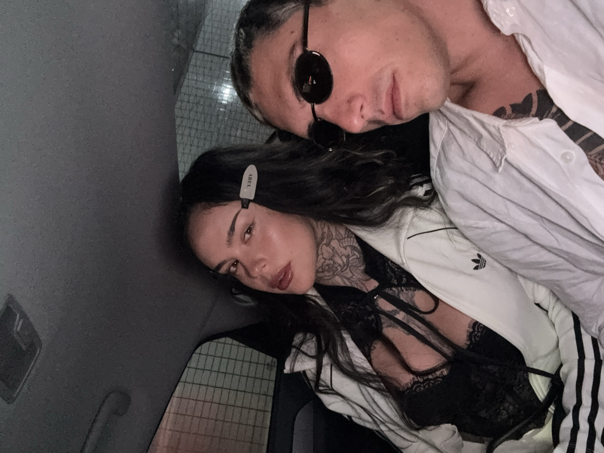 The image depicts a man and a woman posing closely together inside what appears to be a car. The man is wearing sunglasses and a white collared shirt. He has visible tattoos on his neck and chest. The woman is wearing a black lace top under a white Adidas jacket. She also has tattoos on her neck. Her hair is dark and styled with a hair clip that reads "ABEL". The setting suggests a vehicle interior, potentially within an urban environment with a background indicating a possible parking structure or building with a gridded exterior. The location, Seoul, South Korea, cannot be determined from the image content alone.