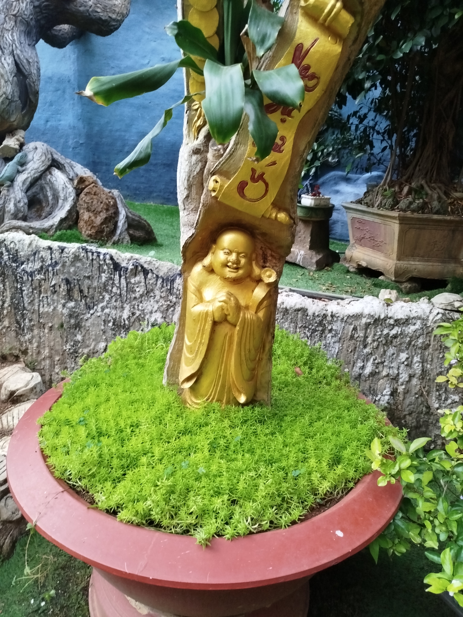 A golden Buddha statue is embedded in a tree trunk inside a red planter filled with bright green foliage. The planter sits in a garden setting in Xuân Thành, Vietnam, with a rock wall and various plants in the background. A golden decorative scroll with Vietnamese writing adorns the tree trunk above the Buddha statue. Other trees and plants are visible, along with a carved rock feature.