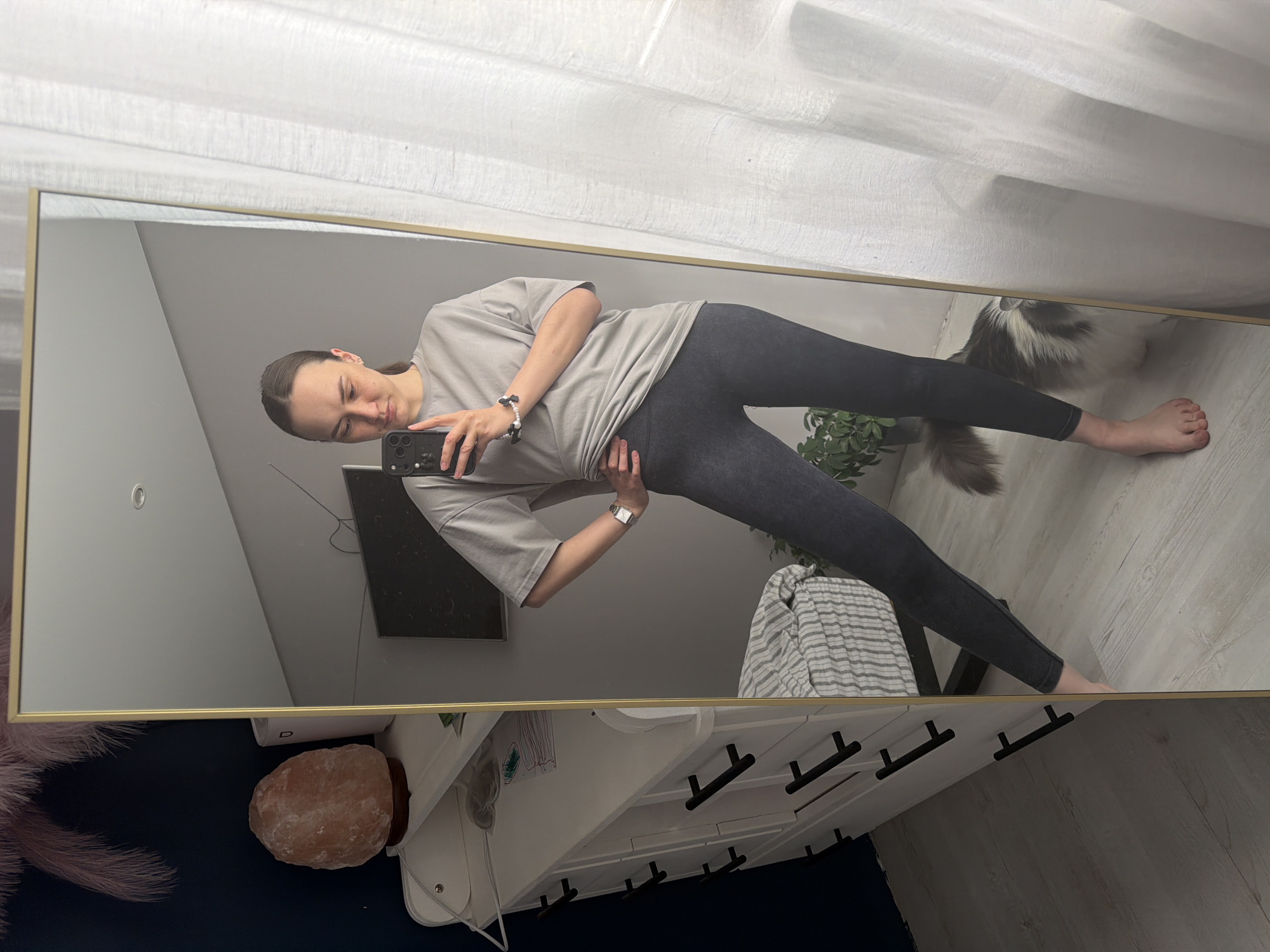 A woman is taking a selfie in a full-length mirror. She is wearing a gray t-shirt and dark leggings. Her hair is pulled back. The mirror reflects a room with a white dresser, a salt lamp, a houseplant, a gray striped throw, and what appears to be a cat's tail. The location of the image is Chaikovsky, Russia.