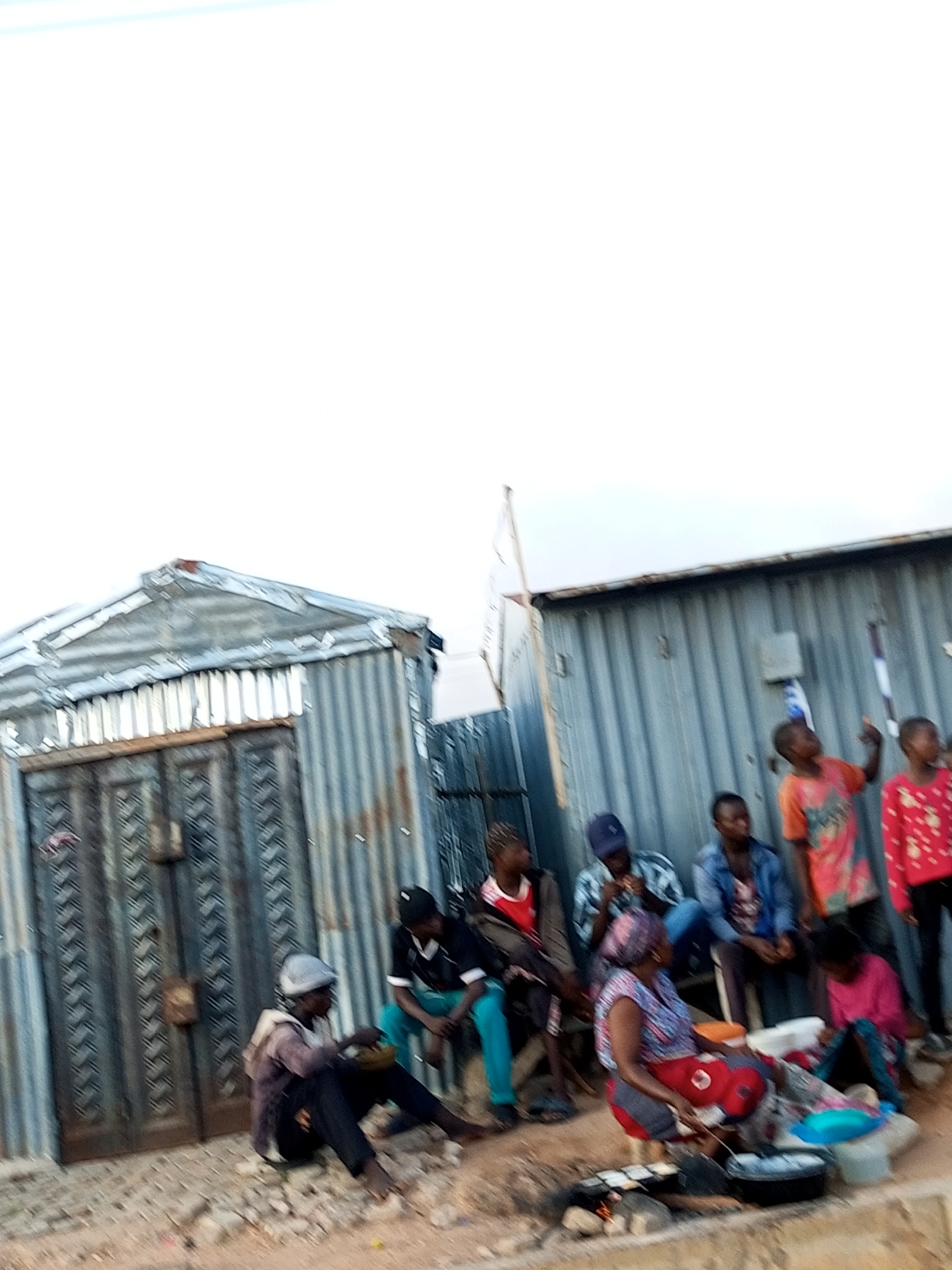 In Tumfure, Nigeria, a group of individuals are gathered outdoors near corrugated metal structures. On the left, a corrugated metal building with large double doors stands. To the right, another corrugated metal structure forms a backdrop. Several people are seated on the ground in front of the latter building. A woman in a patterned top and red skirt is seated in the foreground, attending to cooking on a makeshift stove with pots and food items. Adjacent to her, another person in a black and white top and blue pants is seated. Further to the right, a group of individuals, including children and adults, are standing and sitting. One child in an orange shirt points upwards. The ground is uneven and covered with debris. The sky above is overcast.