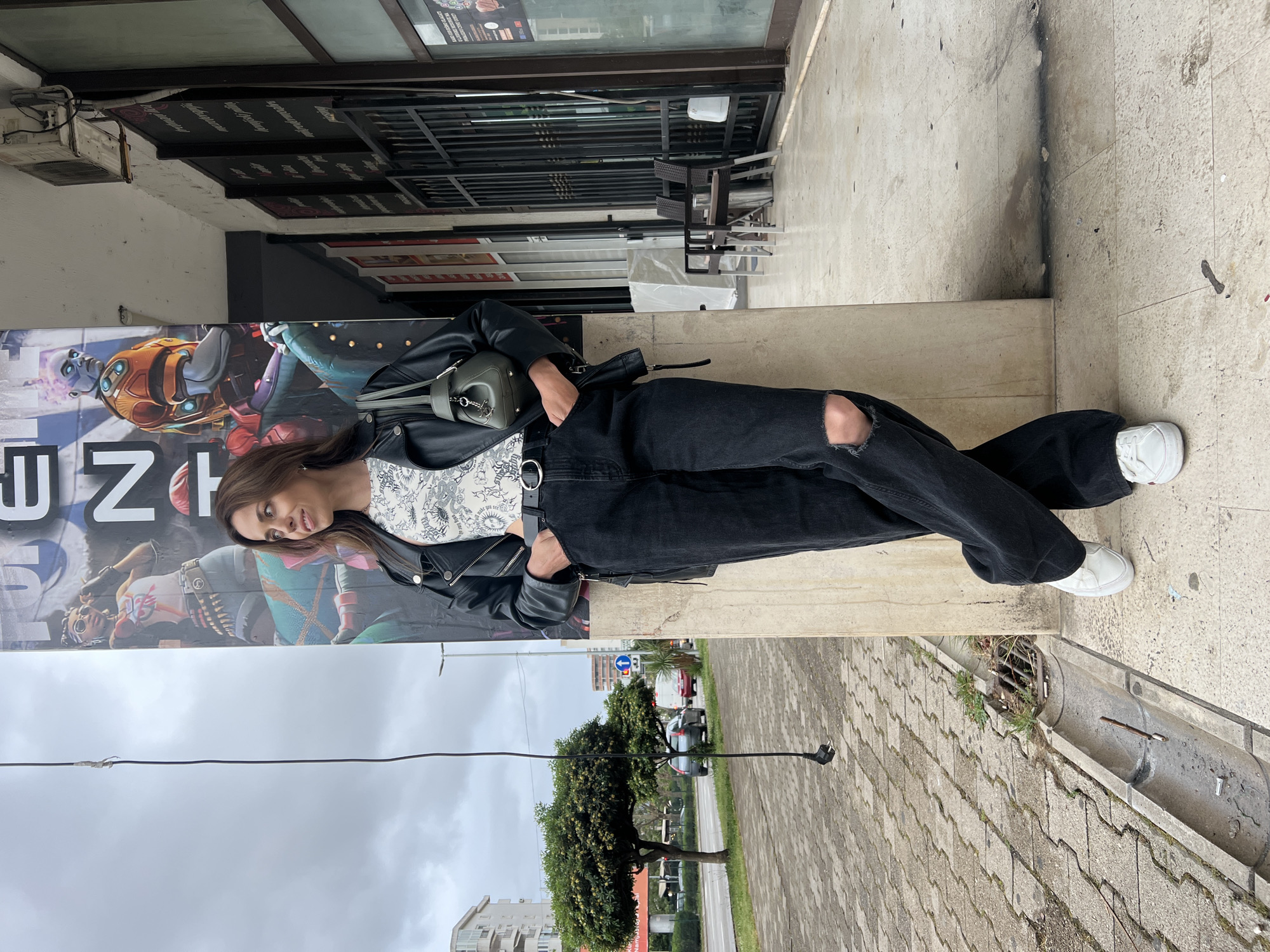 A young woman is sitting on a low concrete ledge in an urban environment. The location is Bar, Montenegro, as evidenced by street signage visible in the background. The woman is wearing a white patterned top, black leather jacket, black jeans with a rip at the knee, and white sneakers. She has a gray purse slung over her shoulder. Behind her is a large advertisement, possibly for a game or entertainment venue, featuring colorful characters. Above the advertisement are signs for a restaurant or business. The weather appears overcast. A power cable runs parallel to the sidewalk on the ground level. The sidewalk is constructed of gray pavers. A storm drain runs parallel to the curb.