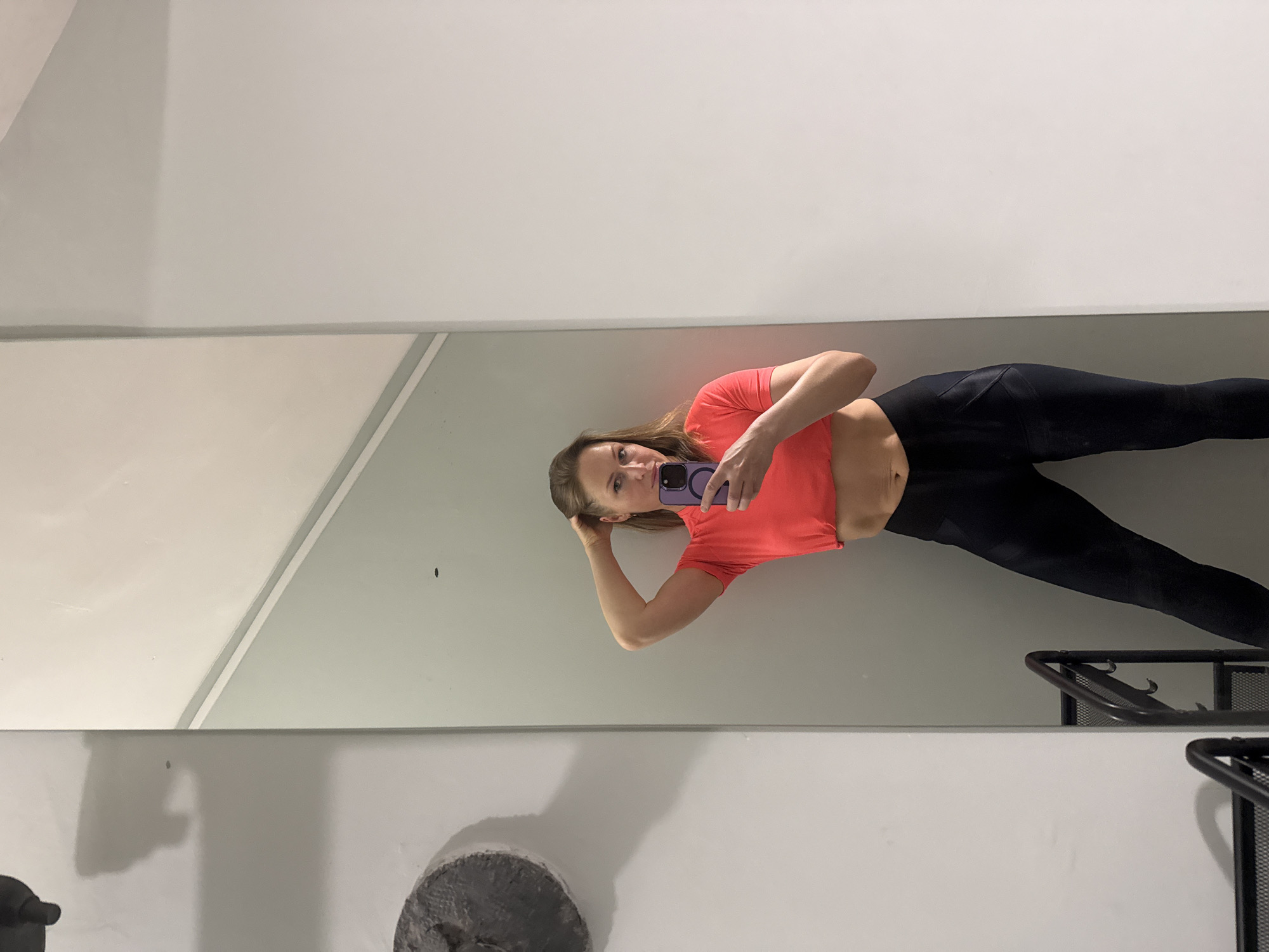 A woman with light brown hair is taking a selfie in a mirror. She is wearing a short-sleeved coral-colored shirt and black leggings. Her right hand is holding a purple phone, and her left hand is resting on her head. The background is a room with white walls and a gray stripe. There is a black metal rack and a gray, textured object (possibly a weight) visible in the lower portion of the mirror reflection. The location is Unknown, Unknown.