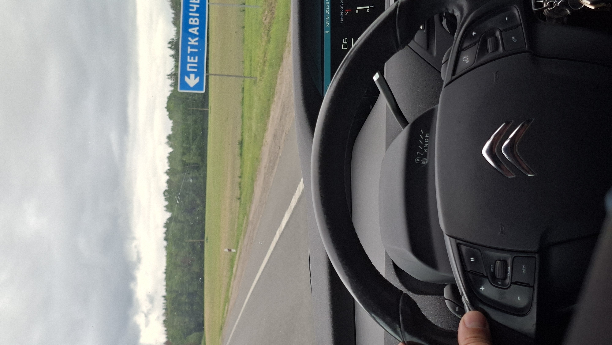The image depicts an interior view from a vehicle, specifically focusing on the steering wheel with the Citroen logo in the center. The vehicle is traveling on a road as seen through the windshield. A road sign indicating "ПЕТКАВІЧЕ" with an arrow pointing left is visible on the roadside. The environment appears to be rural, with fields and trees present. The location is in the city of Дзяржынскі сельскі Савет, Belarus, as indicated by the road sign.