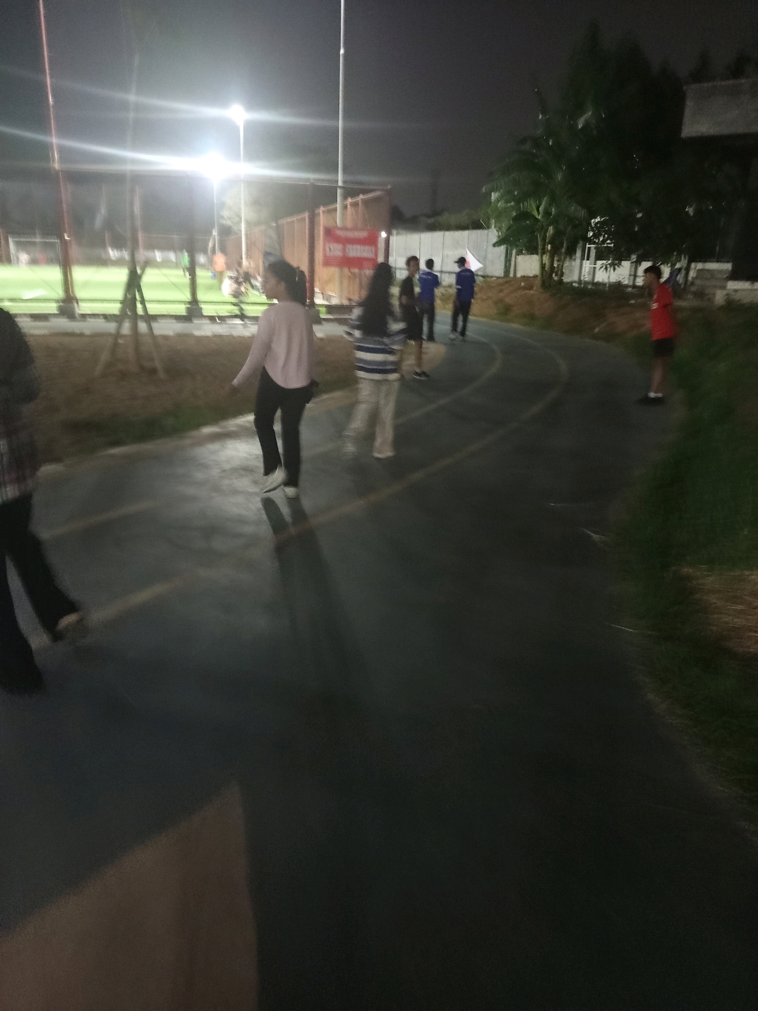 The image depicts an outdoor scene at night, likely a sports facility in West Jakarta, Indonesia. A running track with yellow lane markers is prominent in the foreground, with grass and trees bordering it. Several individuals are visible on the track. In the mid-ground, a person in a light pink top and dark pants walks on the track, casting a shadow. To their right, another person in a striped shirt and light pants walks in the same direction. Further back, on a lit sports field, several figures in athletic wear can be seen, suggesting a game or practice is in progress. The overall lighting comes from overhead lamps, creating a well-lit but somewhat hazy atmosphere typical of night-time outdoor venues.