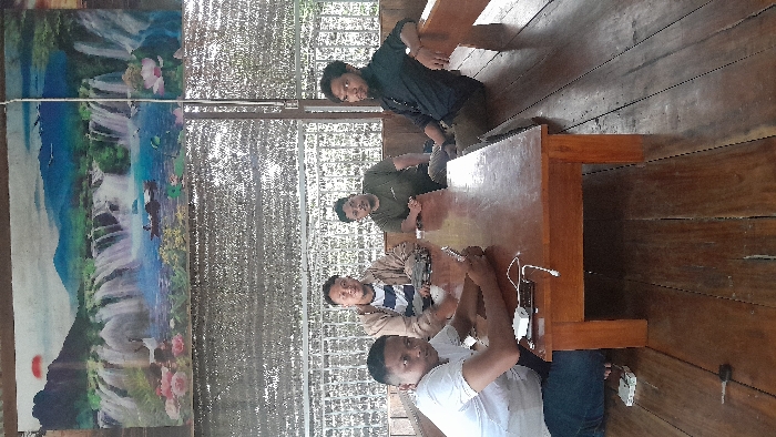 Four adult males are depicted seated around a dark brown rectangular wooden table within an open-sided structure, likely a public eatery, located in the city of Aluebilirayeuk, Indonesia. Above, a mesh tarp serves as a canopy, and a large banner displaying a vibrant landscape with waterfalls, mountains, water, flora, a crane, a boat, and a red sun is affixed to the wall behind them. The floor is constructed from wooden planks.
From left to right:
1. A male in a white t-shirt and blue jeans kneels beside the table, holding a black smartphone and looking down at it. A white power bank connected to a charging cable rests on the table in front of him. A white smartphone or device is on the wooden floor near his left foot.
2. A male wearing a light brown jacket over a striped t-shirt sits on a wooden bench behind the table, resting his right arm on the table's surface and looking towards the camera.
3. A male with glasses, wearing an olive green t-shirt, sits on the wooden bench, with his forearms on the table, looking towards the camera.
4. A male with dark hair and a beard, dressed in a black long-sleeved shirt and dark pants, sits on the wooden bench, leaning back slightly with his right arm on the bench's backrest, and looks towards the camera.
A silver key is visible on the wooden floor near the lower left table leg. Green foliage is visible beyond the structure's open side.