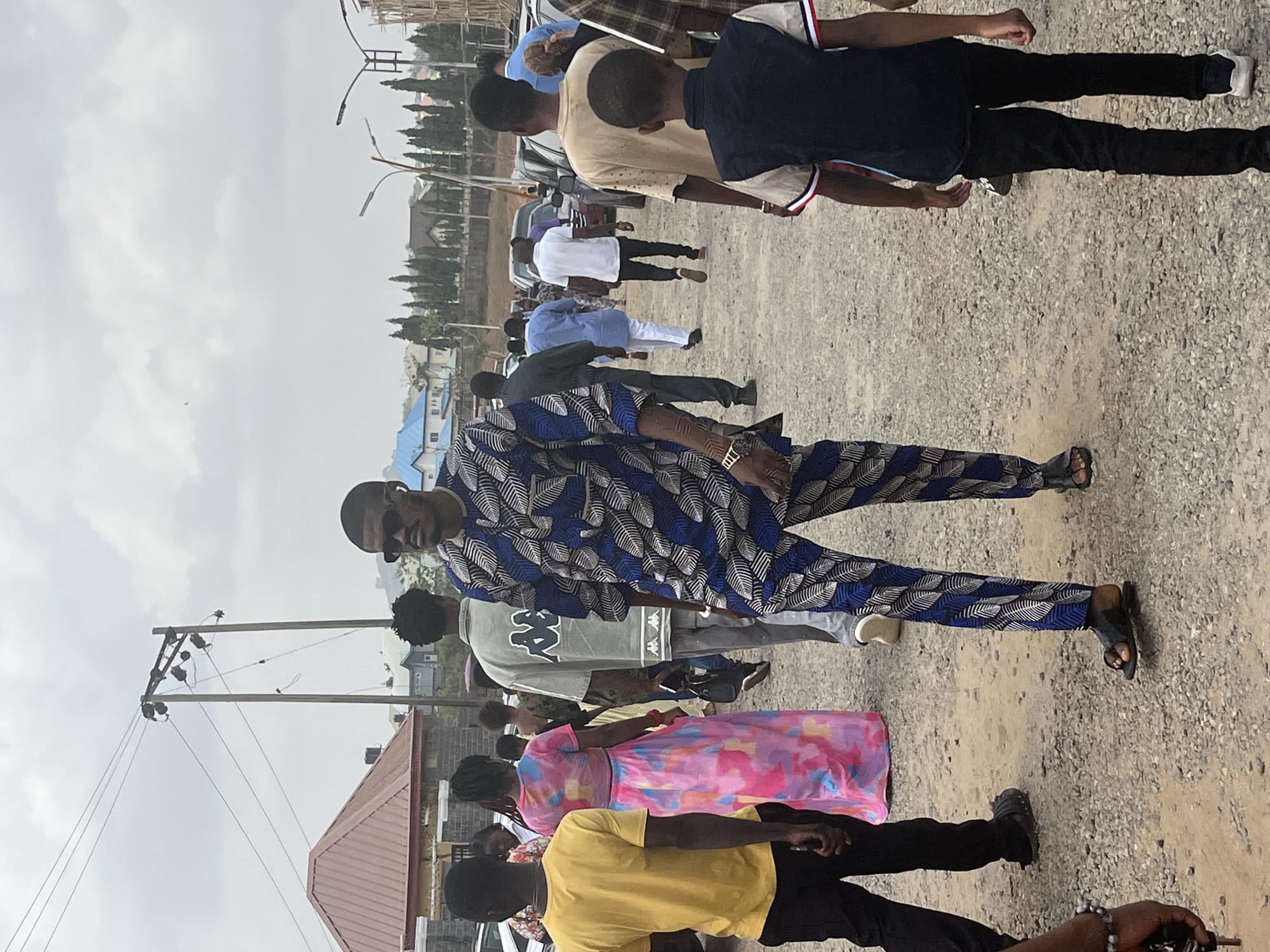 The image captures a group of people outdoors, on what appears to be a gravel-covered area. The scene is likely in a residential or semi-urban part of Abuja, Nigeria, given the presence of buildings, power lines, and trees in the background. The sky is overcast.
The subjects include a man in the foreground wearing a patterned blue and white suit, sunglasses, and sandals. Behind him are other individuals, including a person in a yellow shirt, a person in a pink patterned dress, and others in more casual attire. The people are mostly standing and facing in a general direction, possibly observing something out of frame.
Objects include a building with a red roof, power lines, and assorted clothing worn by the people. Actions include standing and observing. Interactions are not explicitly clear, though the group dynamic suggests a shared purpose or event. The environment is typical of a developing urban area with unpaved ground and basic infrastructure.