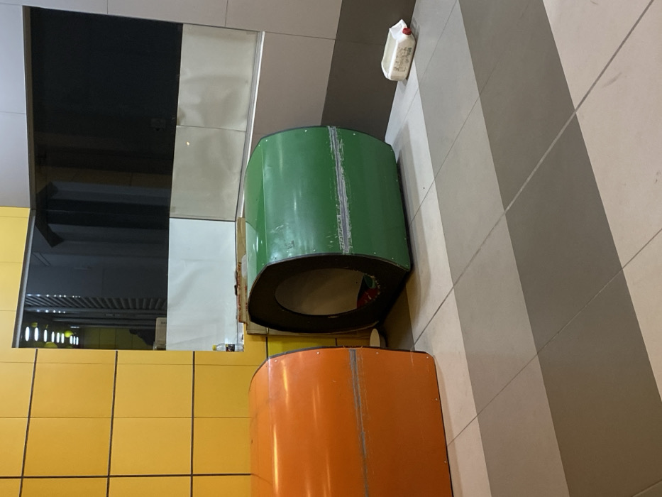 The image depicts an indoor public area within a Mi Teleférico station or concourse in La Paz, Bolivia. The floor is tiled with alternating light and dark gray rectangular panels. Two repurposed Mi Teleférico cable car cabins, disconnected from their tracks, are present. The cabin on the left is orange, showing its rounded exterior. The cabin to its right is green, positioned with its circular opening facing the viewer, revealing red fabric or bedding material inside. Both cabins exhibit surface wear and horizontal seam lines. Between the cabins, a small white bowl rests on the floor. Behind them, a yellow-tiled wall transitions to a dark, reflective window or panel. A small plastic bottle and other items are visible on a low ledge below this window. To the far right, a white plastic jerry can lies on its side on the floor. No individuals are present.