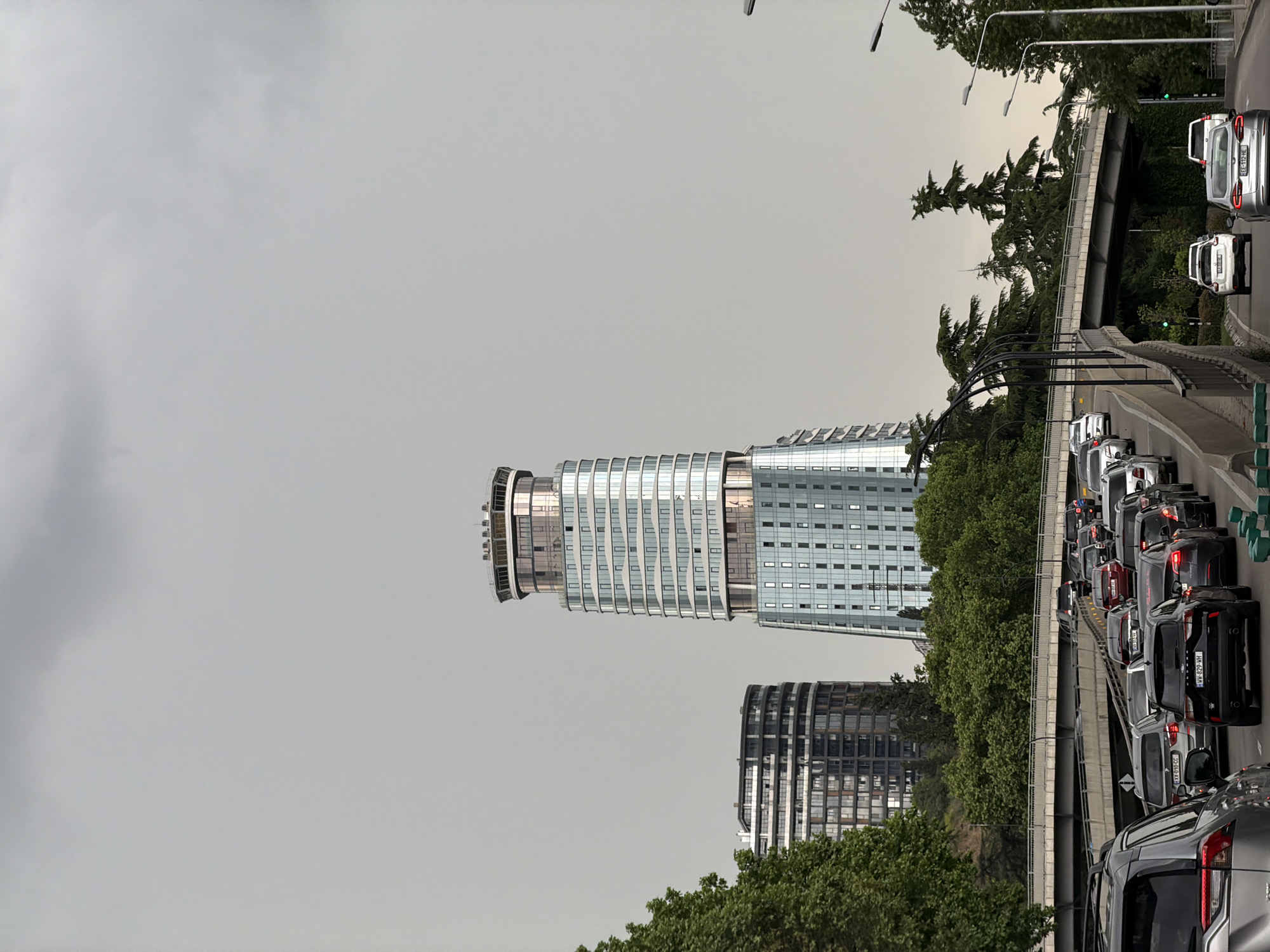 The image captures a modern architectural scene in Tbilisi, Georgia. The sky overhead is overcast, creating a diffused light. Dominating the skyline is a contemporary skyscraper with a unique cylindrical upper portion, surrounded by a sleek, glass-clad facade. Adjacent to this structure is another building, also with a modern design, featuring a darker facade with similar glass elements.
The foreground of the image reveals a busy traffic scene on a bridge or elevated roadway. Vehicles are densely packed, suggesting rush hour or traffic congestion. A line of trees separates the road from the buildings. A bridge or overpass can be seen with several cars on it. The license plate number “XX-010-CC” and “WW-829-WH” is partially visible on the vehicles. In the upper right a car license plate reads “FEE-112-HE.” The scene presents a contrast between urban development and daily life in Tbilisi.