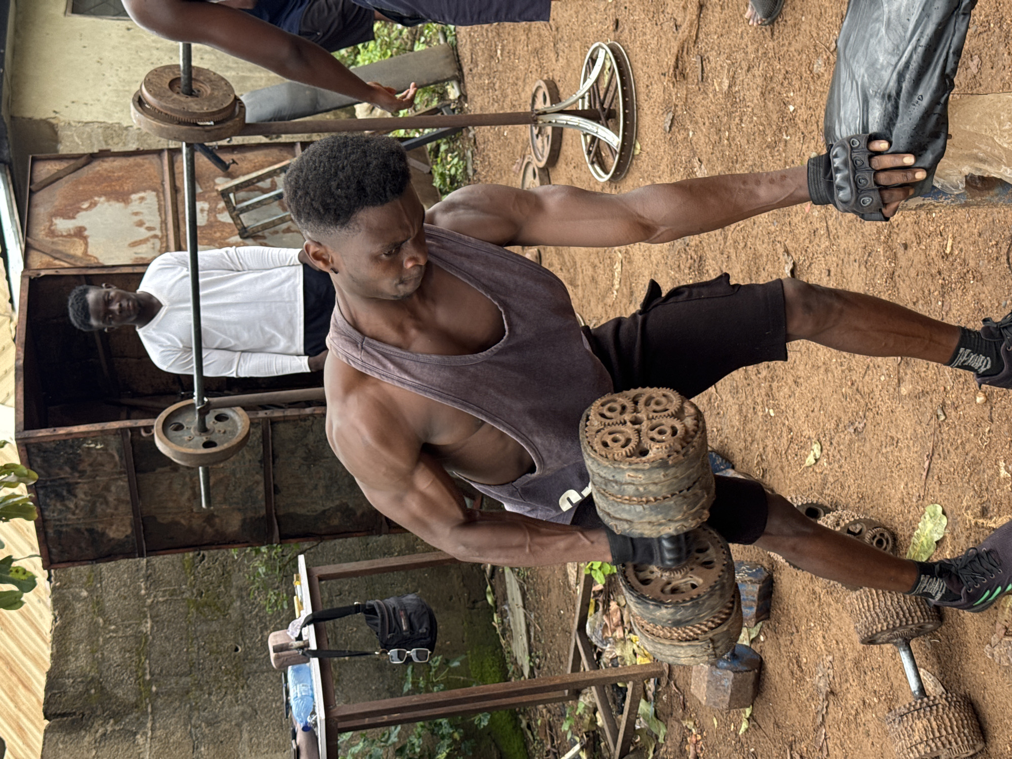 In the city of Akwanga, Nigeria, a muscular Black man in a grey tank top and black shorts is performing a dumbbell row exercise outdoors. He is holding a heavy, rusty, multi-cogged dumbbell in his right hand, with his left hand on a black bag. He wears black workout gloves and black athletic shoes with green accents. Another Black man in a white long-sleeved shirt stands behind him, and a third Black man's arm is visible on the right side of the frame. The workout area is dusty and sandy, with various pieces of weightlifting equipment, including barbells and plates, scattered around. In the background, a corrugated metal structure and a concrete wall suggest a simple, makeshift gym.