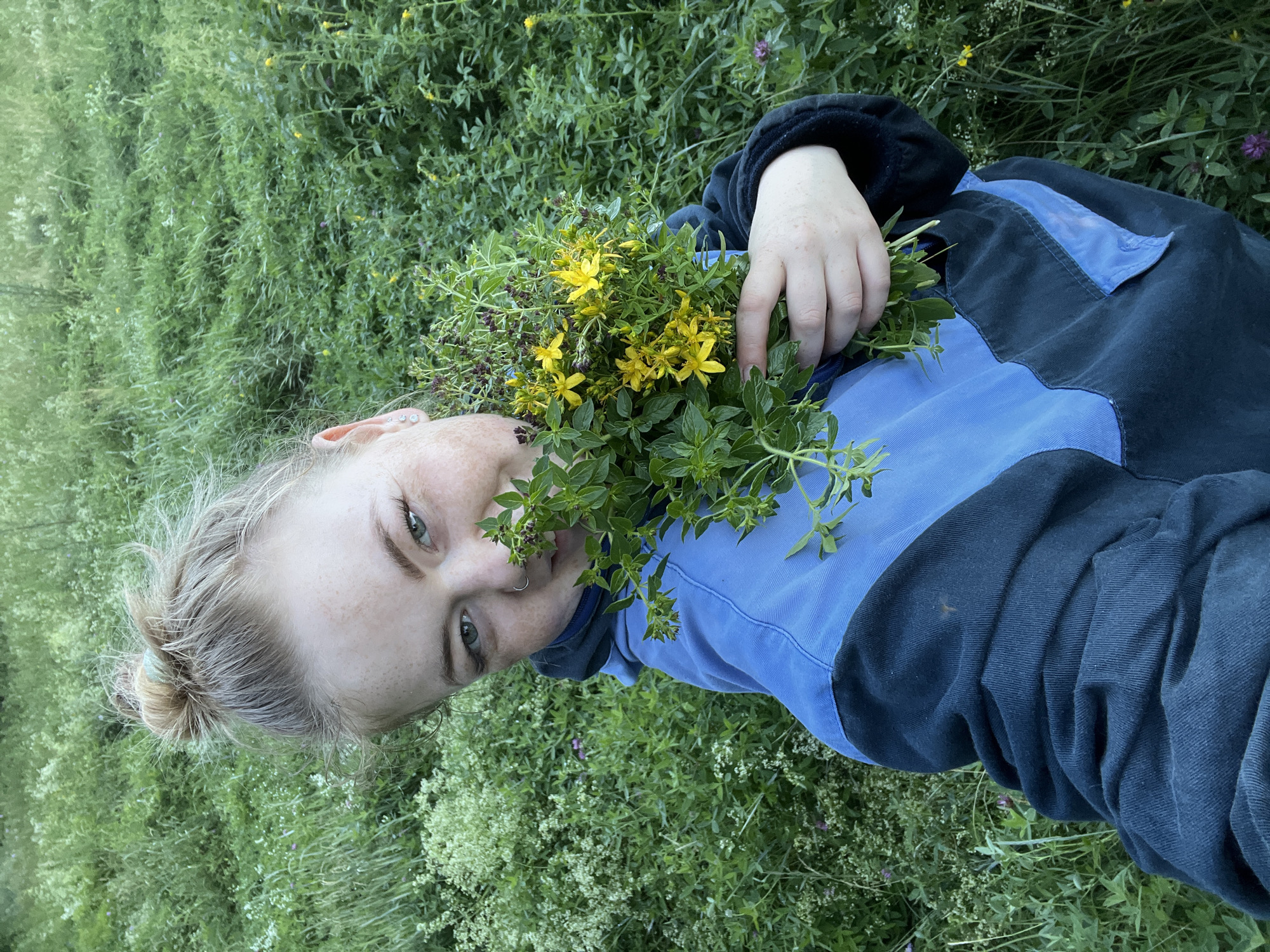 A young woman with blonde hair pulled up in a bun is holding a bouquet of wildflowers, some with yellow petals, in a grassy field. She is wearing a dark blue jacket with light blue accents. Her face is turned towards the camera, and she has freckles and a nose ring. The background is filled with lush green vegetation, including tall grasses and smaller plants. The scene suggests an outdoor, natural environment, possibly a meadow or a field on the outskirts of the village of Olkhovka, Russia, given the presence of common wildflowers. The lighting indicates it is daytime, likely in the late afternoon or early evening based on the softness of the light.