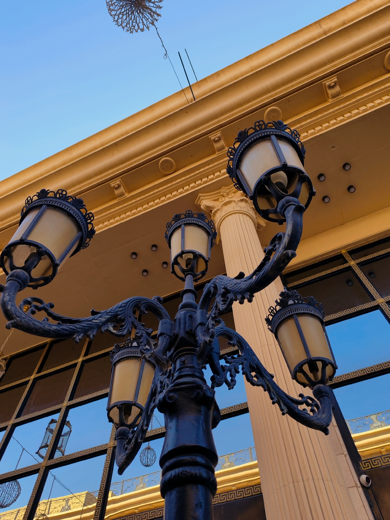 The image shows a low-angle view of an ornate black street lamp with multiple lights in front of a building. The building has a warm yellow facade with classical architectural elements, including fluted columns and decorative moldings. Large windows with a dark grid pattern reflect the blue sky. The street lamp itself is dark metal, with intricate scrollwork and five glass-shaded lamps. One lamp is in the foreground, and four others extend outwards on curved arms. The sky above is a clear, bright blue. The image appears to be taken in an urban setting.