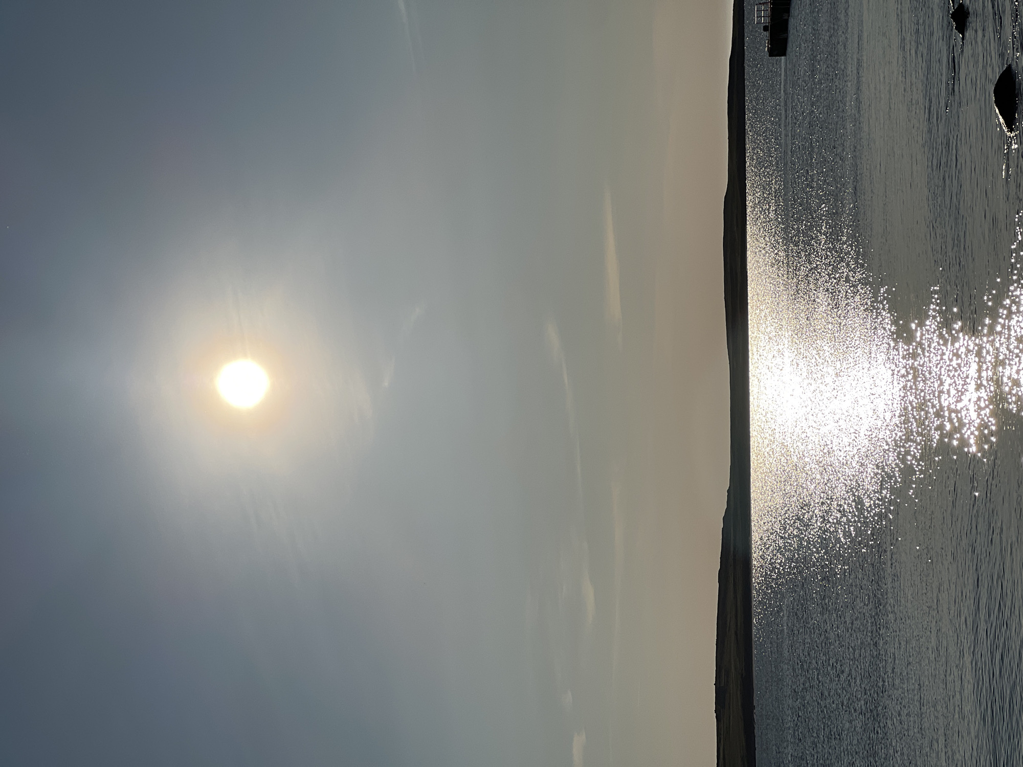 The image shows a wide, calm body of water, likely a lake or bay, reflecting the bright, hazy sun. A dark, low-lying landmass or hill forms the horizon in the distance. The water is textured with shimmering highlights from the sun's glare, creating a glittering effect. In the lower right corner, a dark structure resembling a pier or dock is partially visible. The sky is a pale blue, with a few wispy clouds. The overall impression is one of a serene, natural landscape under a bright, diffused light. The image does not contain any people or animals. The location is given as Lunniaghbeg, Ireland, which implies a coastal or lake environment, consistent with the visual information.