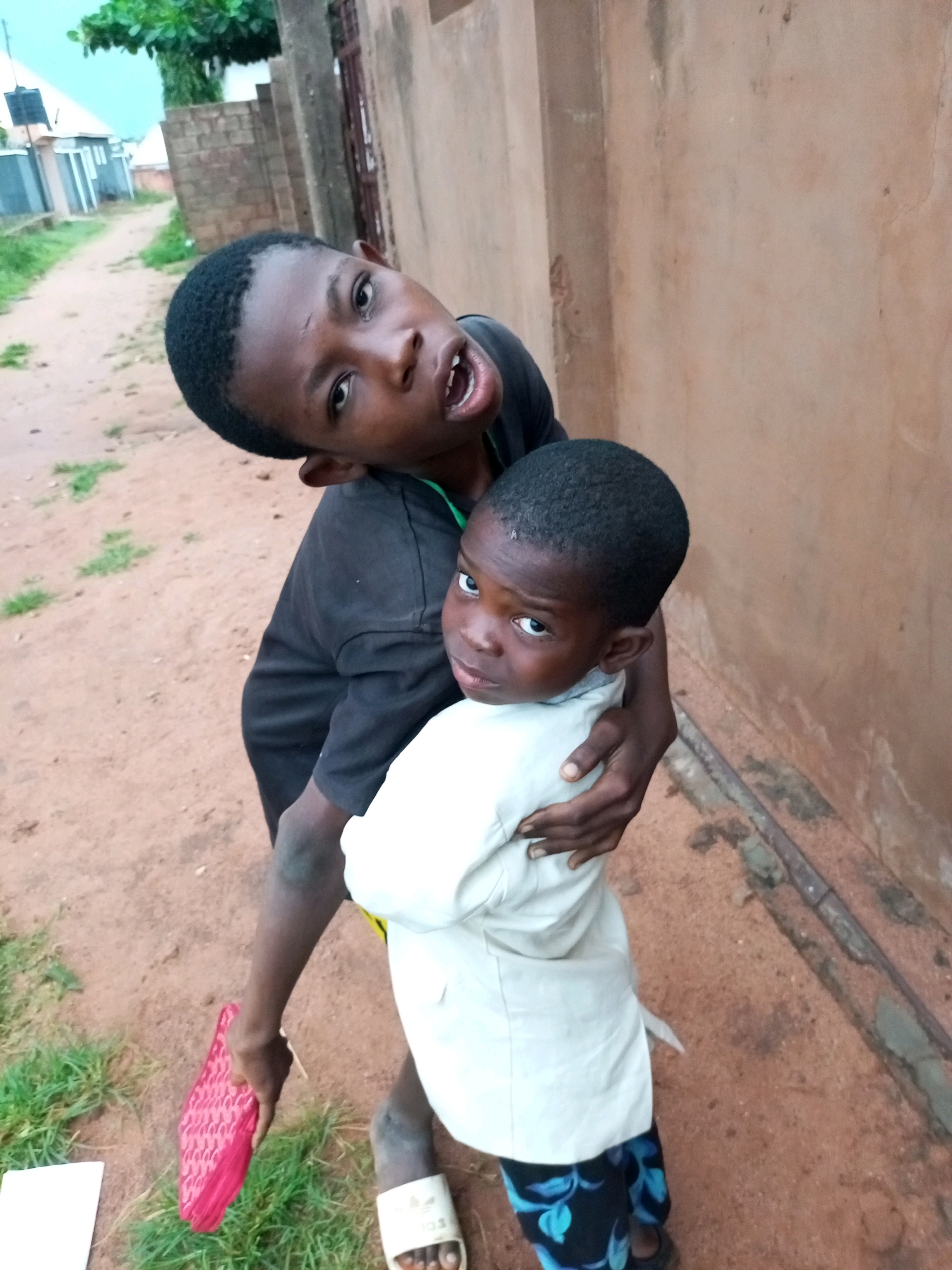In the city of Gombe, Nigeria, two young boys are pictured in an outdoor setting. The older boy, on the left, is wearing a black t-shirt and is holding a younger boy. The younger boy, on the right, is wearing a light-colored shirt and patterned trousers. They are standing on a dirt path next to a brown wall. The background shows a dusty road with some green grass and buildings. The older boy is leaning back and looking towards the camera with his mouth open. The younger boy is looking towards the camera with a slightly worried expression. The older boy is holding a pink, woven object in his left hand.