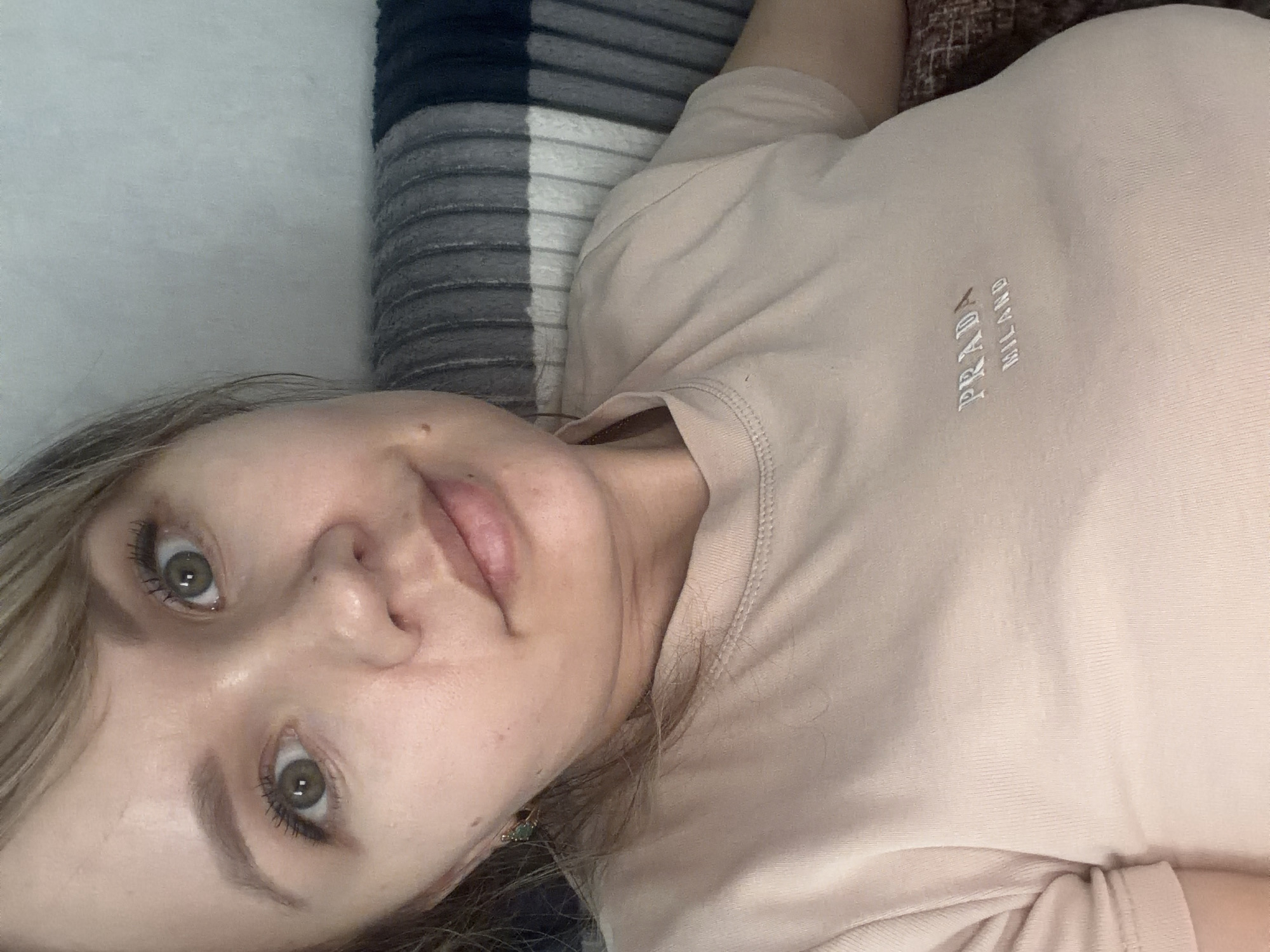 The image captures an overhead view of a person, likely female, lying down indoors. Her head is tilted to the left, and she looks directly into the camera with a neutral, relaxed expression. She has light-colored hair framing her face, and her prominent green or hazel eyes are accented with visible mascara. Defined eyebrows and a small mole on her left cheek are noticeable features. A small, dark earring can be seen on her left ear.
She is wearing a light beige or pale pink short-sleeved t-shirt. The brand name "PRADA MILANO" is clearly displayed in silver or white lettering on the chest, oriented vertically. The fabric of the t-shirt appears soft and comfortable.
The background consists of a striped blanket or bedspread, featuring distinct bands of dark blue, gray, and white, suggesting a comfortable indoor setting like a bed or sofa. The lighting is soft and diffused, creating an even illumination across the scene without harsh shadows. The overall impression is one of a casual, intimate moment captured in an indoor environment.