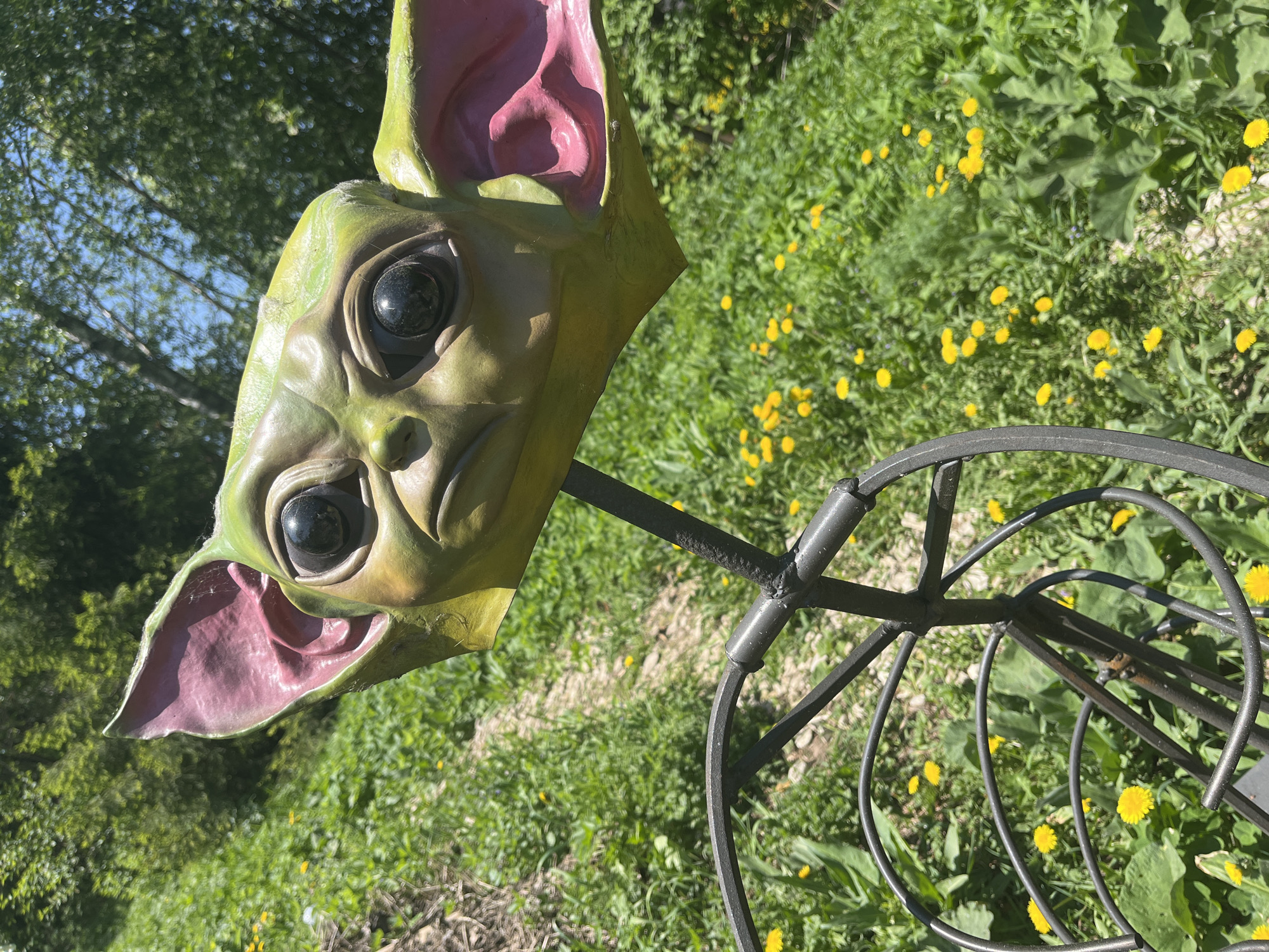 A close-up shot depicts a green-painted mask with exaggerated features, including large eyes and ears colored pink on the inside, affixed to a dark metal stand with a circular design. The mask appears to be outdoors, set against a backdrop of grass and numerous yellow flowers, suggesting a lawn or meadow. The setting is partially shaded, with trees visible in the upper left corner. The scene's location is Unknown, Unknown.