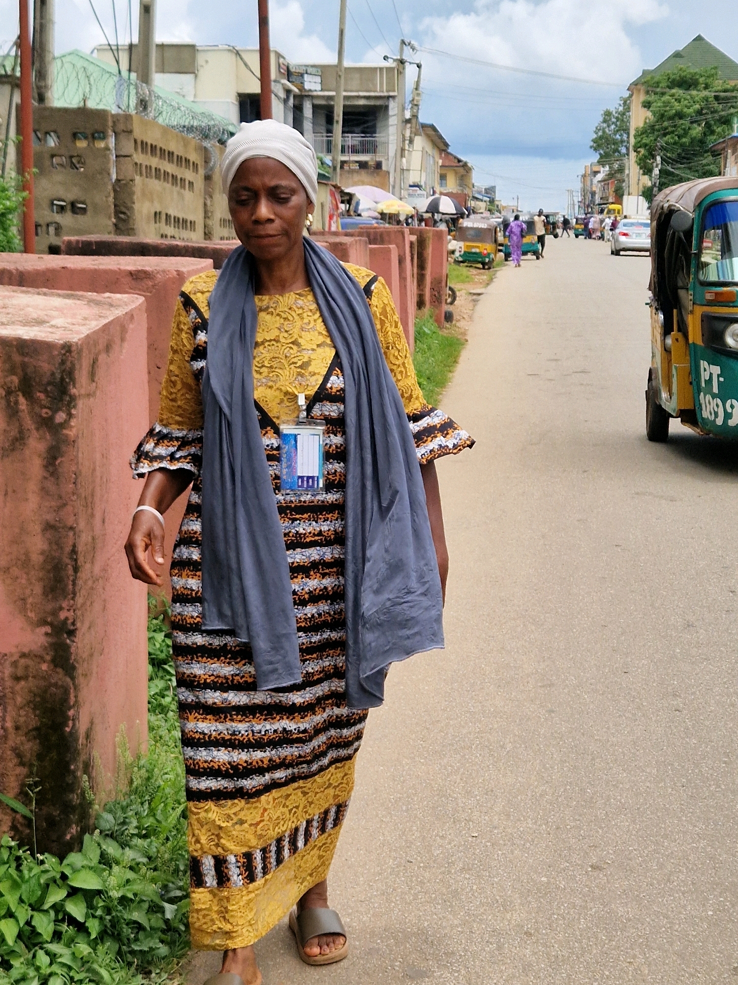 A woman wearing a yellow lace top, a grey scarf, and a striped dress walks on a paved street. She has a white headwrap and a white bracelet on her left wrist. A blue and white ID badge hangs from her neck. The street is lined with buildings and a green-roofed structure. In the background, several auto-rickshaws and a car are visible. The scene is likely captured in Jos, Nigeria.