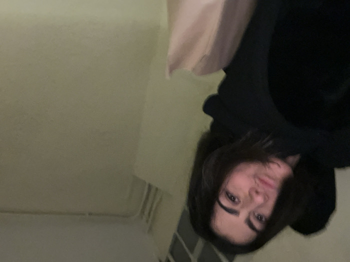 A selfie-style image, rotated, features a person with dark hair and brows, fair skin, and a black hooded sweatshirt. The individual is looking at the camera with a slight smile. A portion of a white sheet or curtain is visible above the person's head. The background consists of a light green wall and what appears to be a white radiator or similar fixture. The lighting is soft and muted.