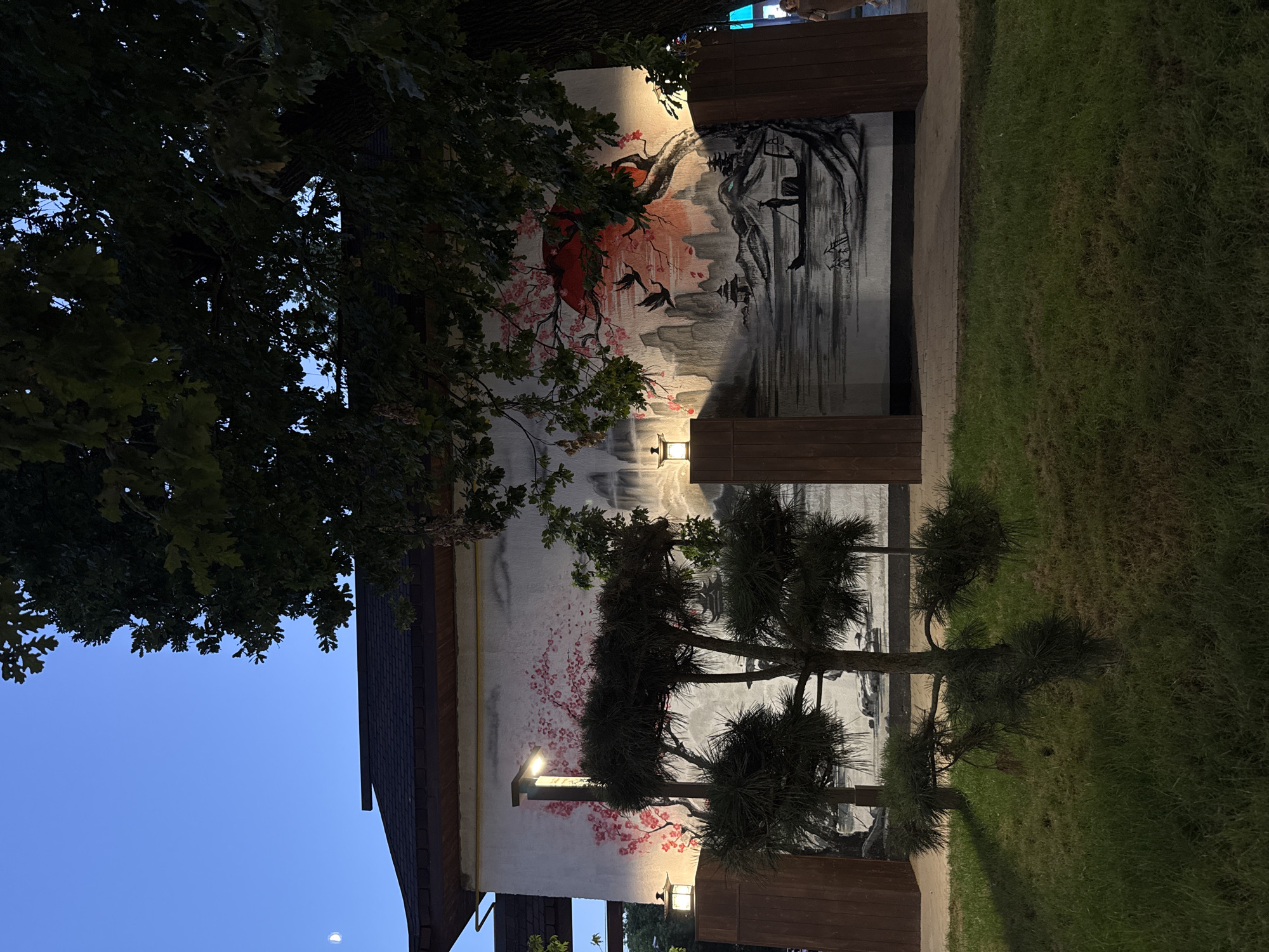 The image depicts a building with a mural painted on its exterior wall. The mural is in a traditional East Asian style, featuring a landscape with mountains, water, a boat with a figure, and a large red sun. Cherry blossoms are also depicted in pink. The building has a dark roof with exposed wooden beams and is illuminated by two outdoor lamps. In front of the mural, there are several small, sculpted pine trees and a grassy area. To the right of the mural, a large tree with dark green foliage partially obscures the view. The sky above is a deep blue, suggesting it is dusk or twilight, with a sliver of the moon visible. A person is visible walking on a paved pathway to the far right of the image. The location is identified as Shymkent, Kazakhstan.