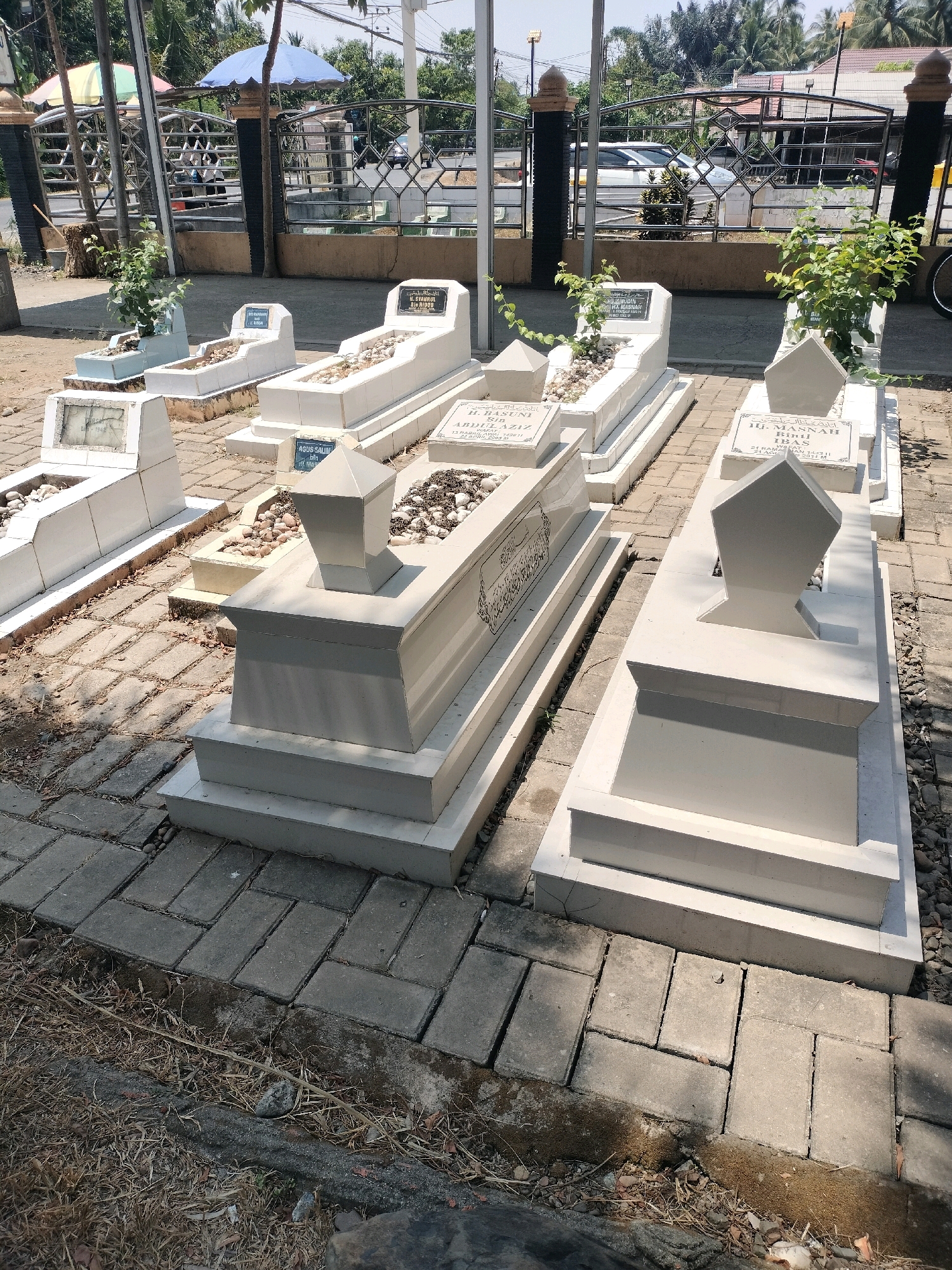 The image depicts a section of a cemetery in Sungai Raya Utara, Indonesia, characterized by a series of white, modern gravestones arranged on a paved walkway. The gravestones are rectangular with tiered bases and some feature decorative finials. The ground is covered with grey interlocking brick pavers, and some areas outside the paved section are filled with dry grass and dirt. In the background, a metal fence encloses the cemetery, with a street and vehicles visible beyond it. Trees and buildings are also part of the wider environment. The lighting suggests it is daytime with sunlight casting shadows. There are no people visible in the immediate vicinity of the graves. Some gravestones have inscriptions, but they are not clearly legible in the provided image. The overall scene is one of order and preservation within a cemetery setting.