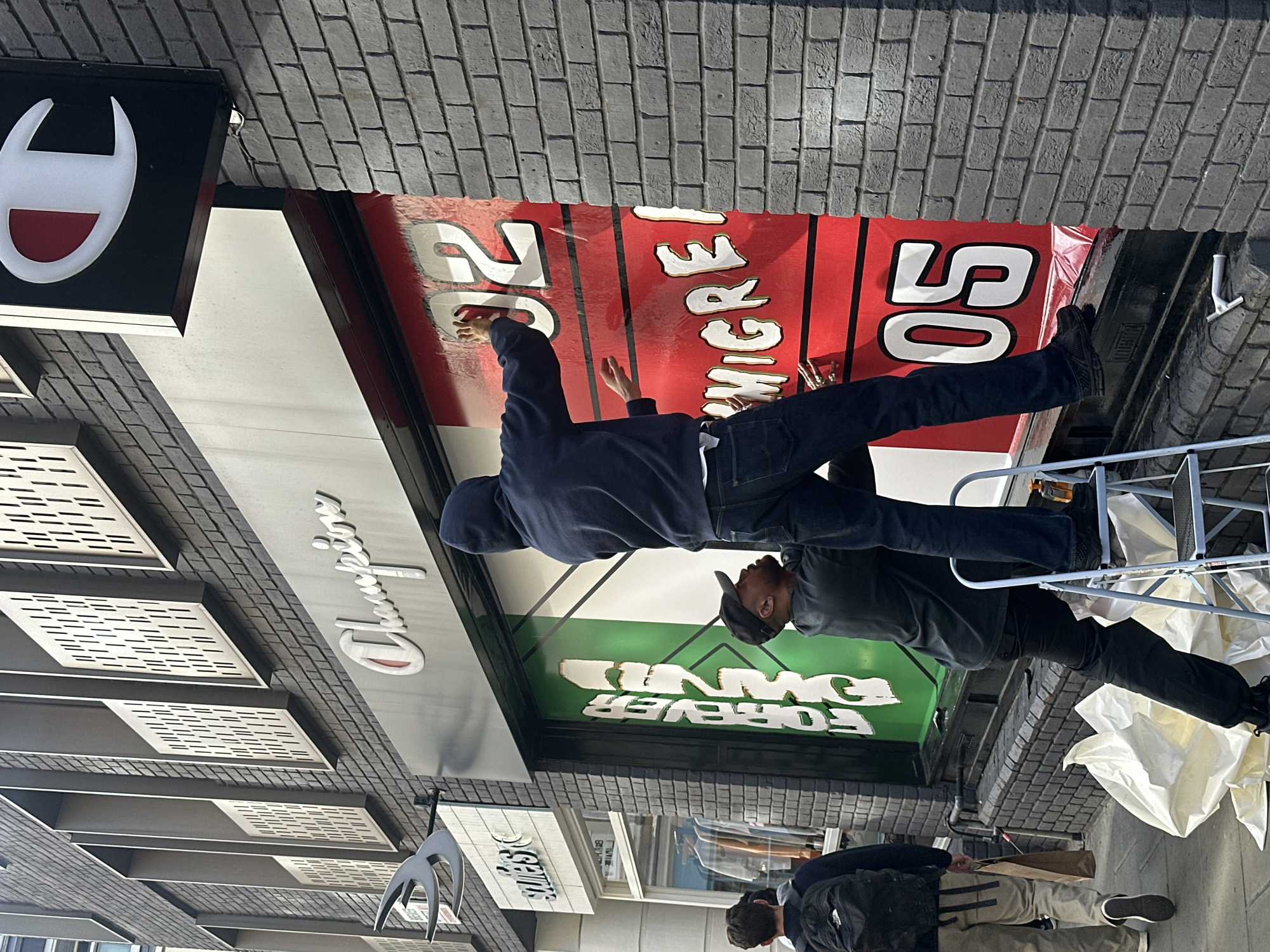 This image captures an outdoor urban scene in London, United Kingdom, during a bright day, showing two men engaged in installing or adjusting a large storefront banner. A third individual is partially visible in the lower foreground.
The central activity features a man, dressed in a dark hoodie and jeans, standing on a silver stepladder. He is facing away from the viewer and reaching up to smooth out or apply a large red banner, with his hands positioned near the bold white number '20'. To his right, another man, wearing dark attire, is also on a stepladder, with only his lower body and leg visible next to a large white number '50' on the same red banner. White protective tarps are spread across the ground near the base of the stepladders, indicating a work in progress.
The storefront is prominently branded with a black sign featuring the iconic white and red 'Champion' logo (stylized 'C' shape). Below this, the word 'Champion' is written in script on a grey panel. The large red banner displays white text with yellow outlines, including the numbers '20' and '50', and partially visible words like 'WHERE'. Vertical black lines are also present on the red banner, possibly mimicking a sports field. Below the red section, a green banner extends across the storefront, bearing the text 'FOREVER OWNED'. A smaller, partially obscured sign further down appears to show the word 'STUSSY'. The building's facade is constructed from dark bricks, with additional modern white slatted panels on the upper left. The overall scene depicts commercial street maintenance or a display setup.