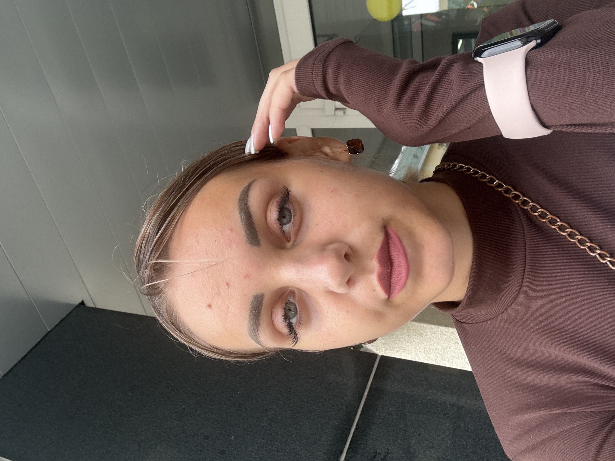 A young woman with light brown hair pulled back, exhibiting acne on her forehead, poses for a selfie. She wears a brown turtleneck shirt with a gold chain necklace. Her left hand is raised to her head, and her right arm is bent, displaying a light pink smartwatch. She has on long false eyelashes and dark lipstick. She is outdoors, with a dark grey wall on the left and a building with windows and a yellow balloon visible in the background. The scene is likely located in Yuzhno-Sakhalinsk, Russia, given the presence of the yellow balloon which is often associated with celebrations.