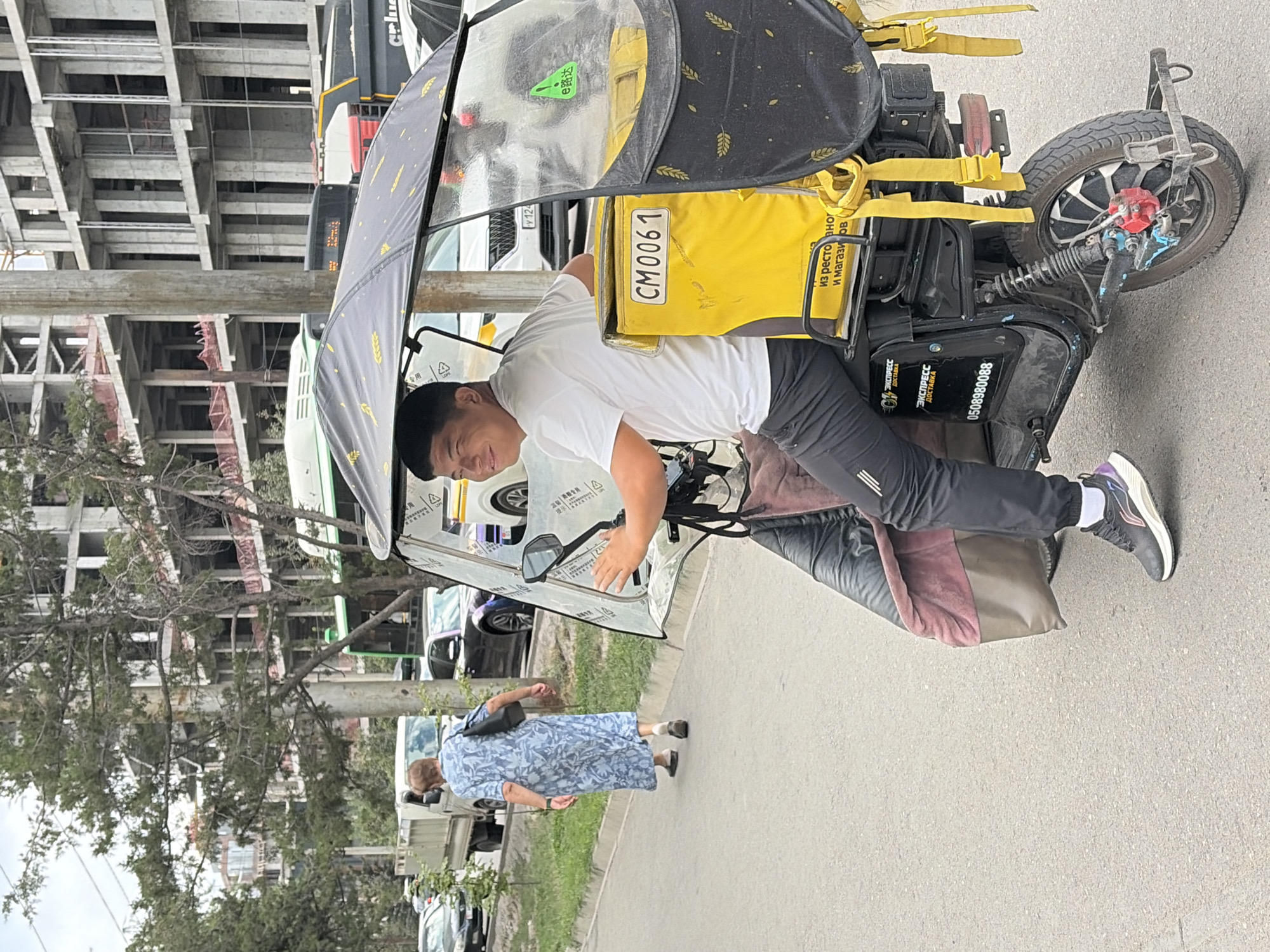 In this medium shot, a young Asian man, wearing a white t-shirt and black athletic pants with a stripe pattern, smiles at the camera as he sits on a black electric scooter. The scooter has a large yellow insulated delivery bag attached to the back with "CM0061" written on a white plate. The bag is adorned with a black cover patterned with yellow wheat stalks and has yellow straps. The scooter also features a transparent canopy for protection from the elements. Behind the scooter, a woman in a blue floral dress with a black bag over her shoulder walks away from the viewer on a paved road. In the background, an unfinished concrete building stands tall, suggesting a construction site. Several vehicles, including a white truck, a black car, and a bus, are visible on the road, indicating an urban setting. The overall scene depicts a street in an unknown city, with the courier on his scooter being the primary subject.