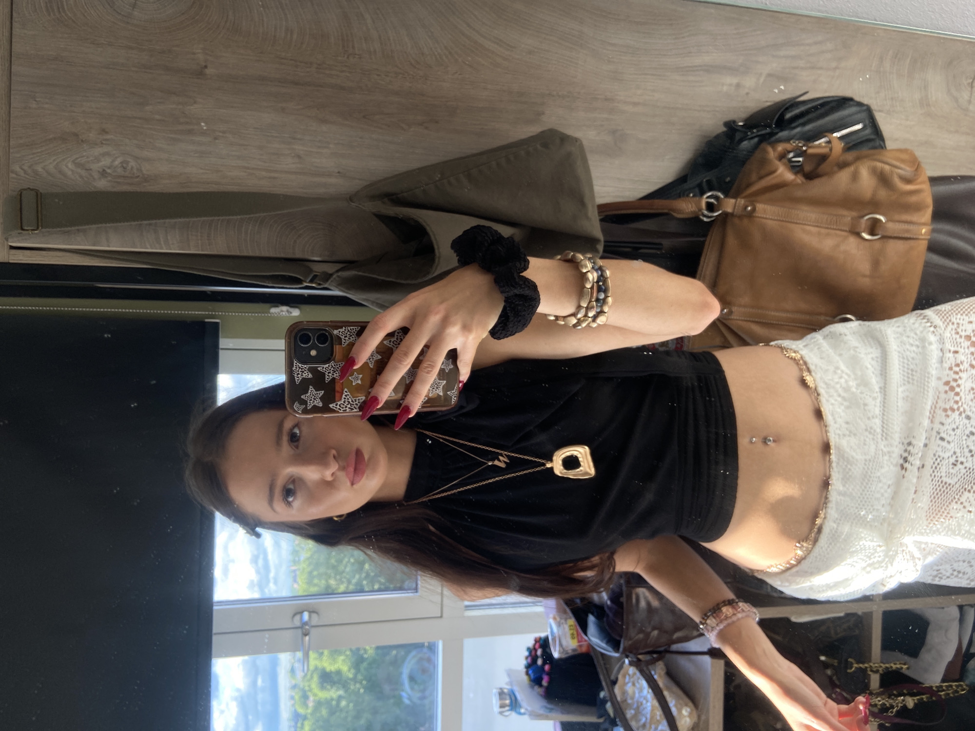 A young woman with long, dark hair takes a selfie in a mirror. She is wearing a black halter top, a white lace skirt, and several necklaces and bracelets. She also has a belly button piercing. In the background, there is a window with a view of trees and buildings, and a wardrobe with several handbags hanging on it. The room appears to be a bedroom or dressing area. There is no identifiable location information for London, United Kingdom.
